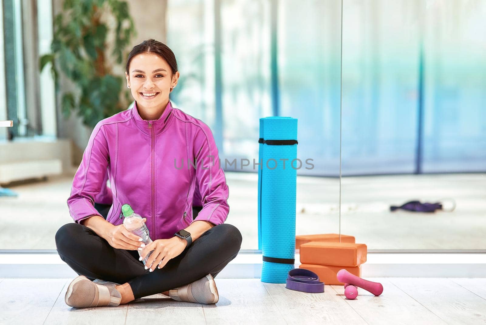 Young beautiful woman in sportswear sitting on the floor against the mirror wall in studio, looking at camera and smiling. Fitness equipment on the wooden floor by friendsstock