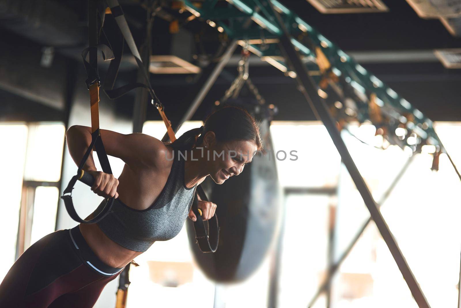 Side view of young beautiful fitness woman in sport wear doing TRX workout at industrial gym, doing push ups. Sport, training, wellness and healthy lifestyle