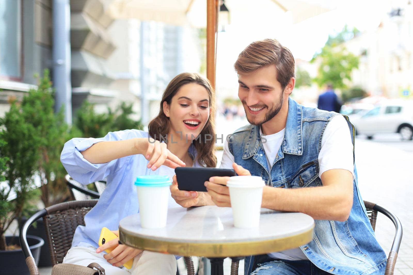 Attractive young couple in love drinking coffee while sitting at the cafe table outdoors, using mobile phone