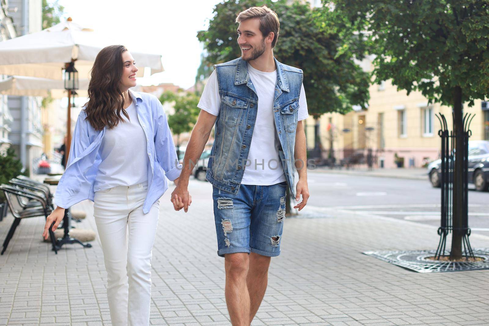 Image of lovely happy couple in summer clothes smiling and holding hands together while walking through city street