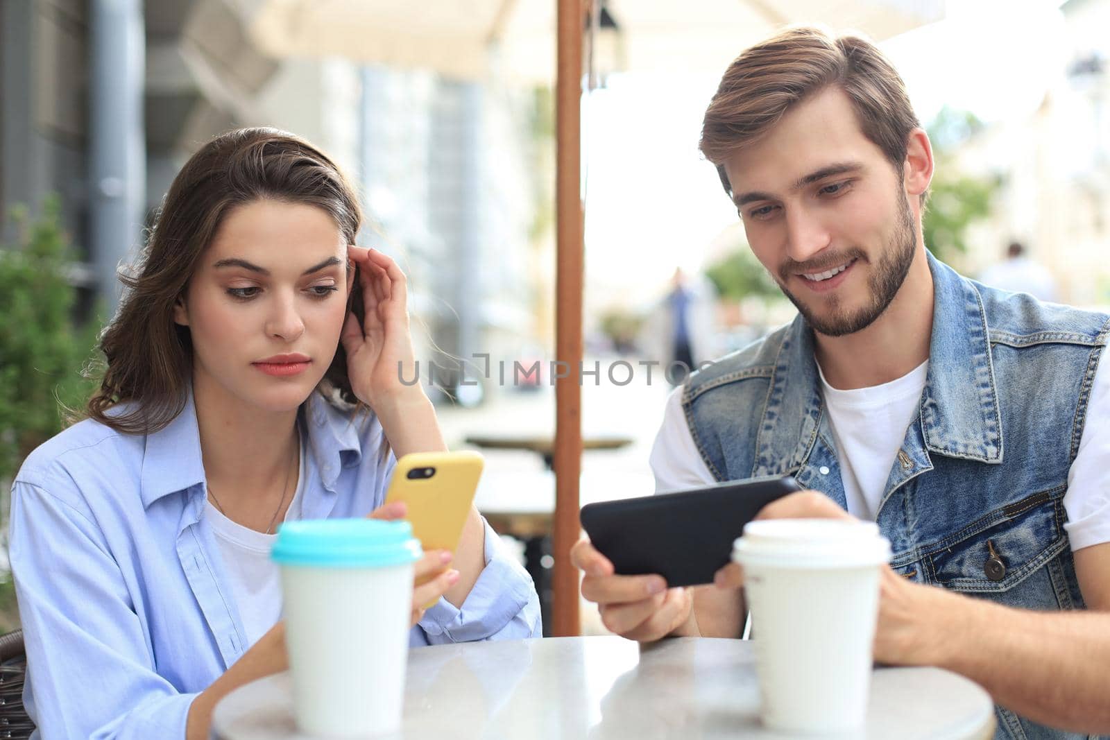 Attractive young couple in love drinking coffee while sitting at the cafe table outdoors, using mobile phone