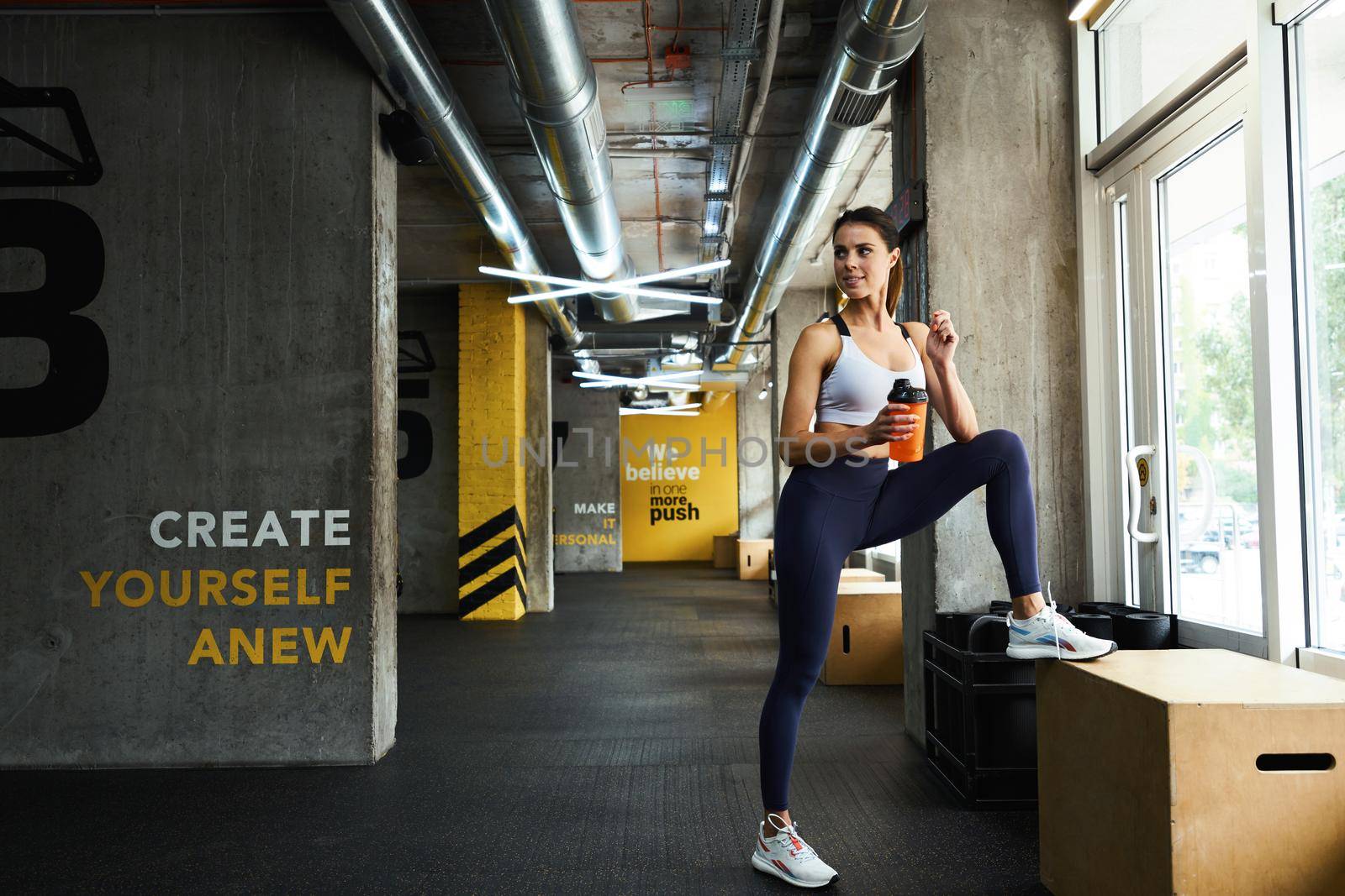 Resting after workout. Full length of a young beautiful fitness woman in sportswear holding bottle of water and looking aside while standing near window at gym. Sport, wellness and healthy lifestyle
