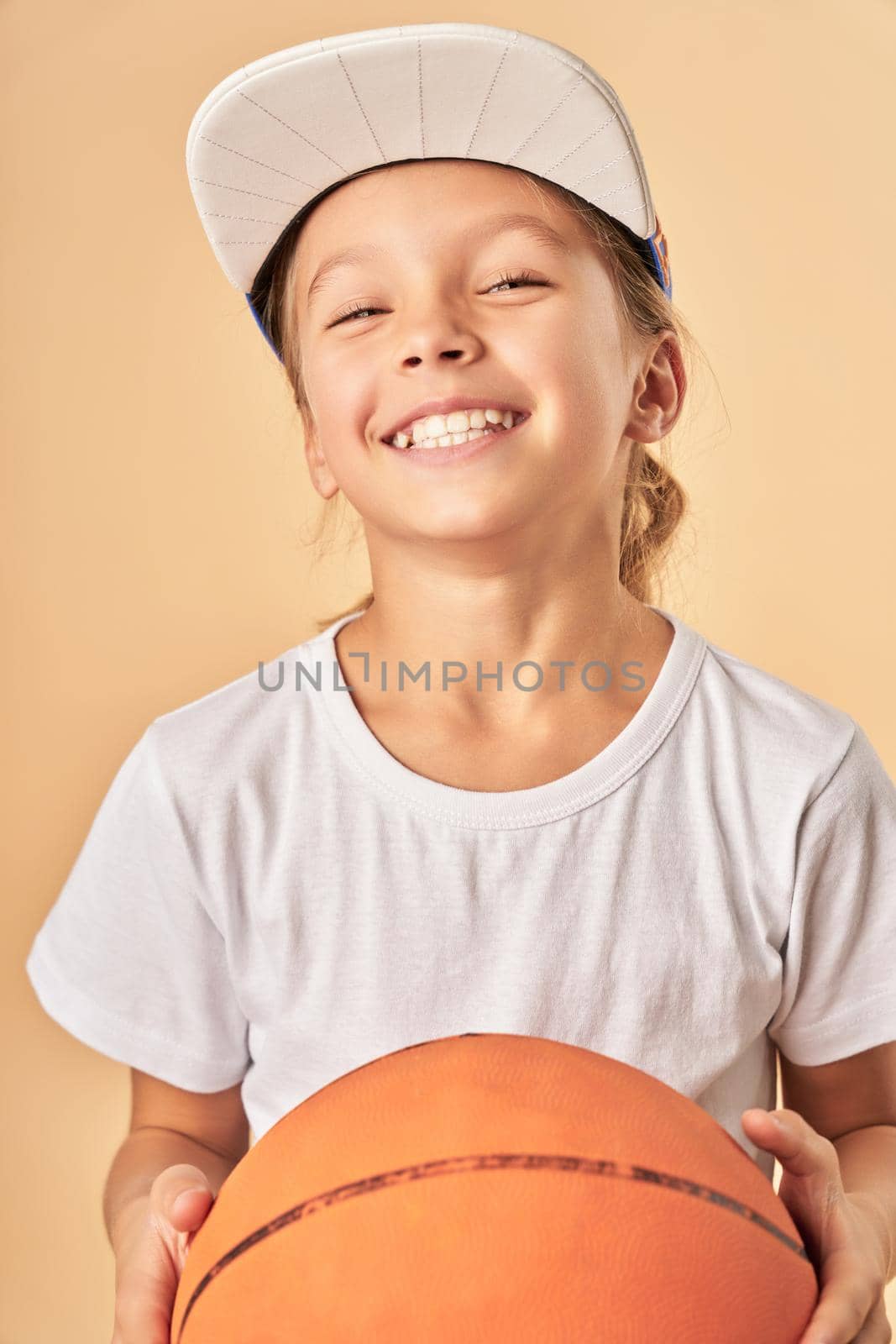 Adorable girl basketball player looking at camera and smiling while standing against light orange background