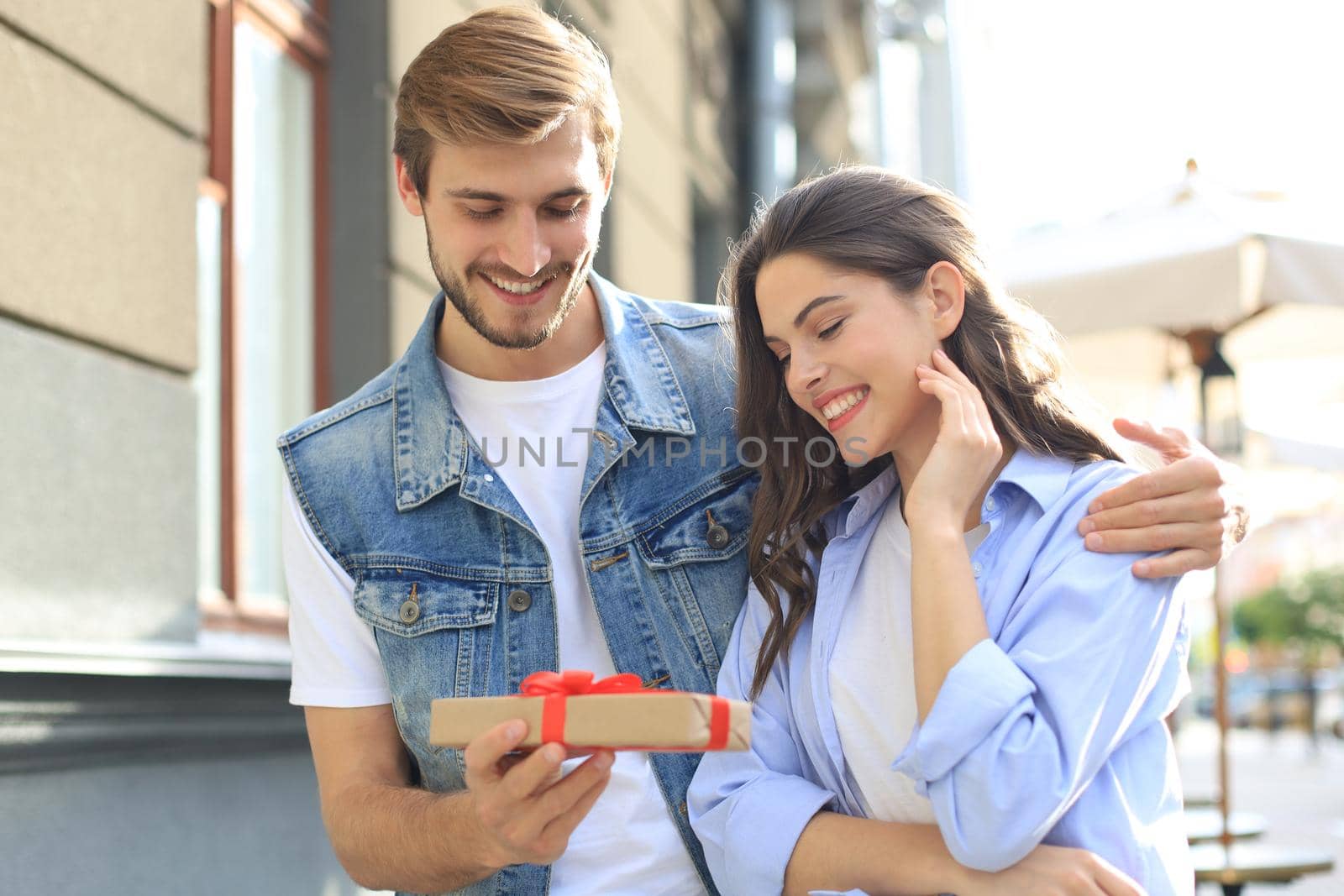 Astonished excited couple in summer clothes smiling and holding present box together while standing on city street