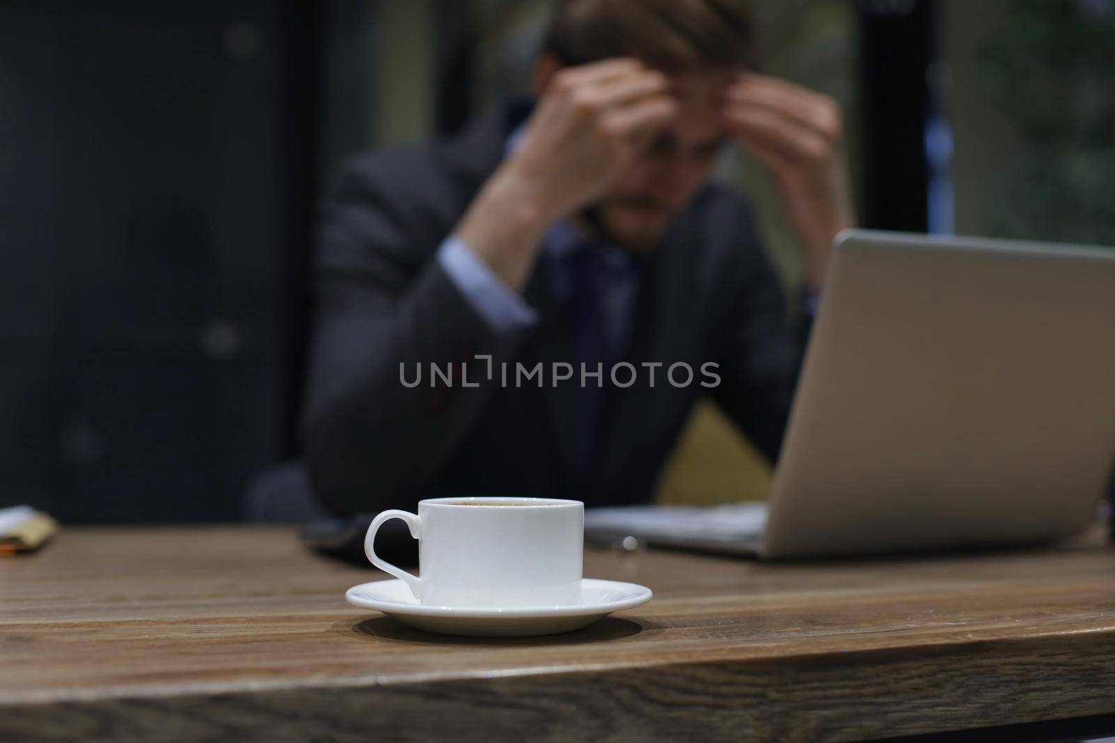 Image of tired young business man in suit keeping face in hands while sitting in the office
