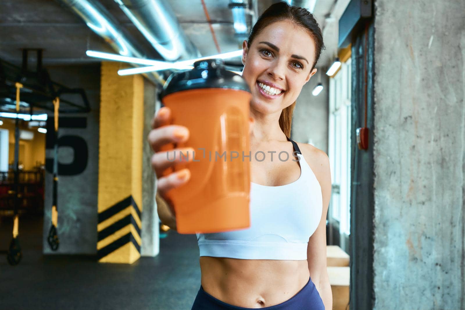 Portrait of a young beautiful and cheerful fitness woman in sportswear holding bottle of water and smiling at camera while exercising at gym. Sport, wellness and healthy lifestyle