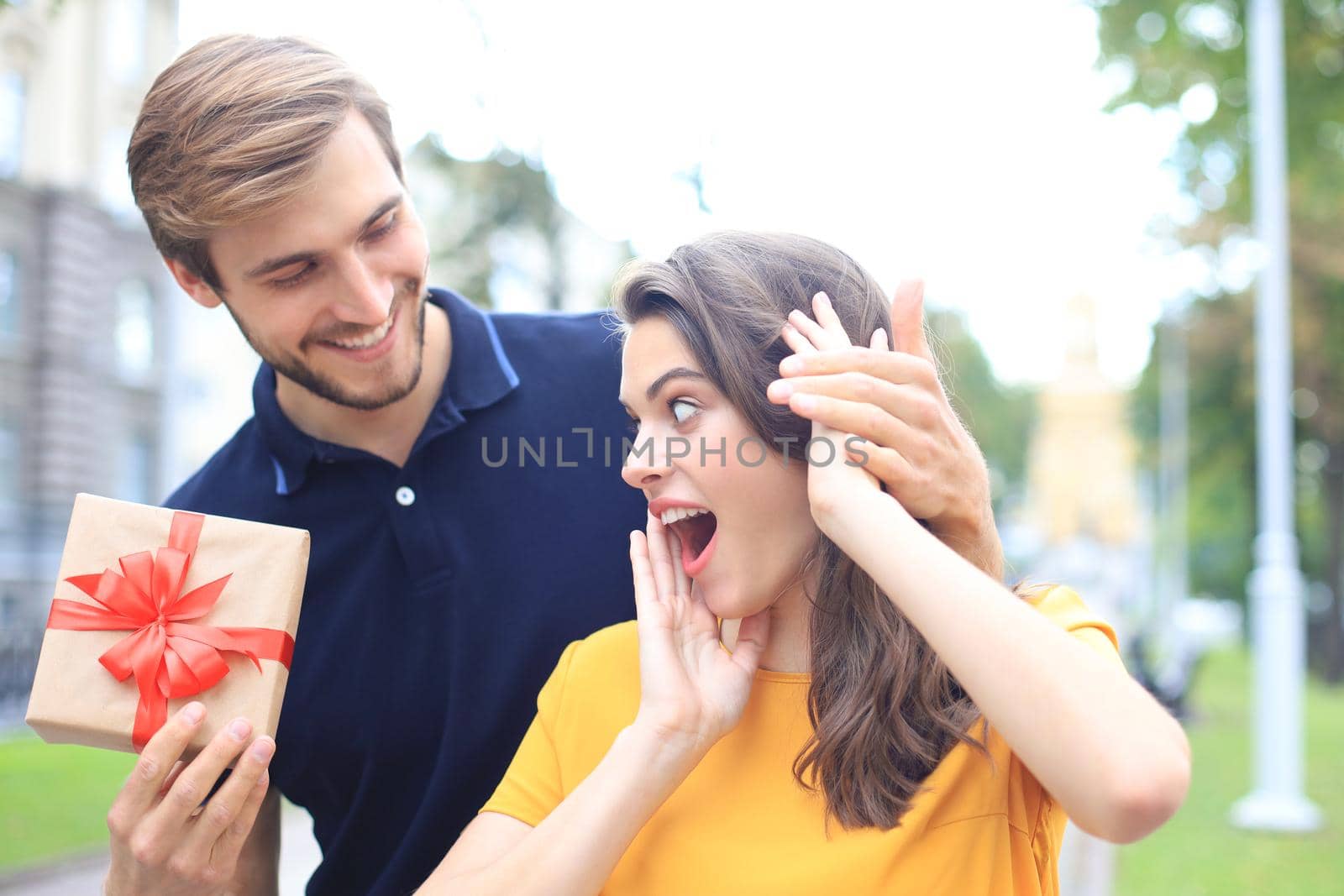 Astonished excited couple in summer clothes smiling and holding present box together while standing on city street