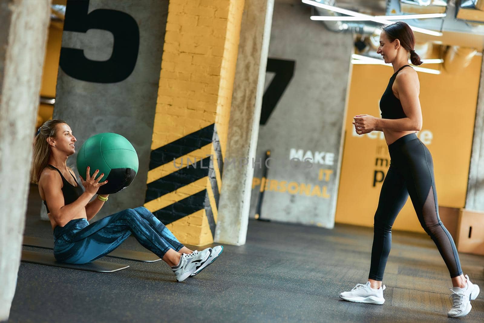 Young athletic woman in sportswear exercising with fitness ball at gym with assistance of personal trainer, doing abs exercises by friendsstock