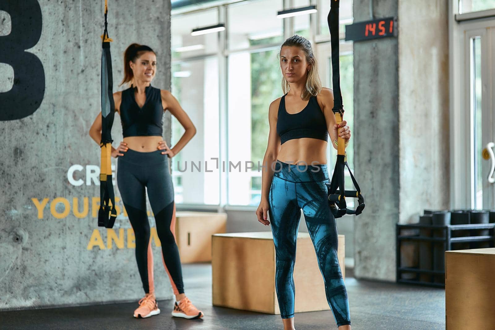 Two young beautiful fitness women looking at camera while exercising with trx fitness straps at gym. Sport, partner workout, wellness and healthy lifestyle