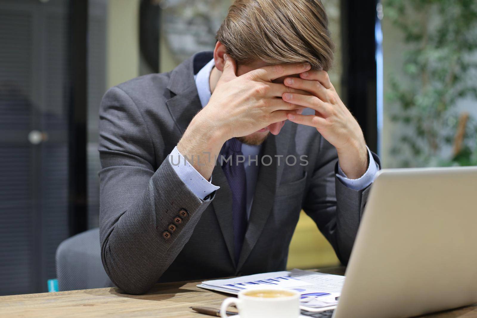 Image of tired young business man in suit keeping face in hands while sitting in the office