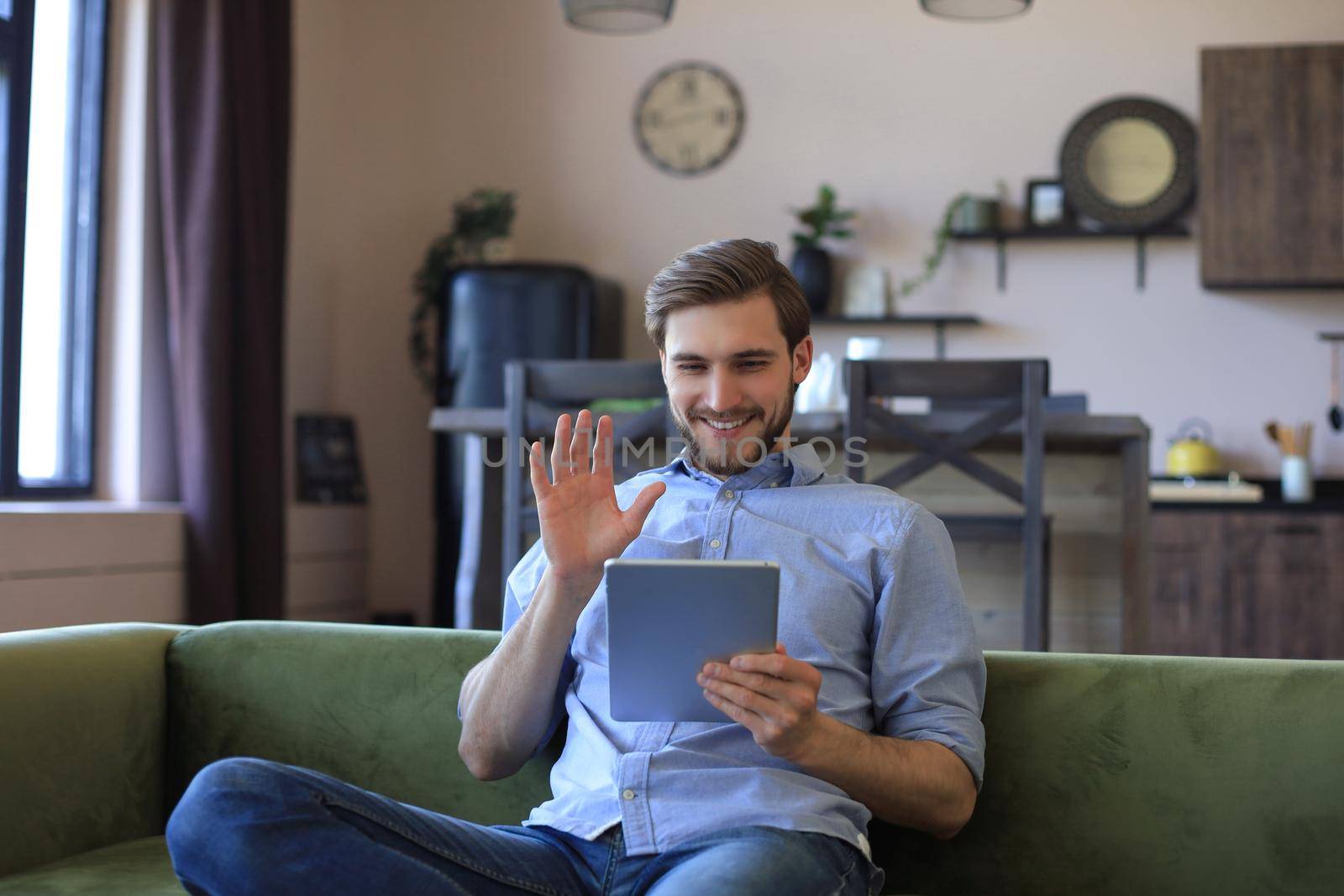 Smiling handsome man holding and using tablet with social networks at home, happy freelancer making online shopping, sitting on cozy sofa at living room