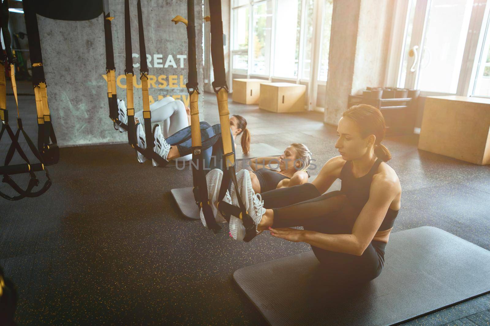 Working out together. Three young athletic women in sportswear sitting on yoga mats at gym and exercising with trx fitness straps. Sport, workout, wellness and healthy lifestyle