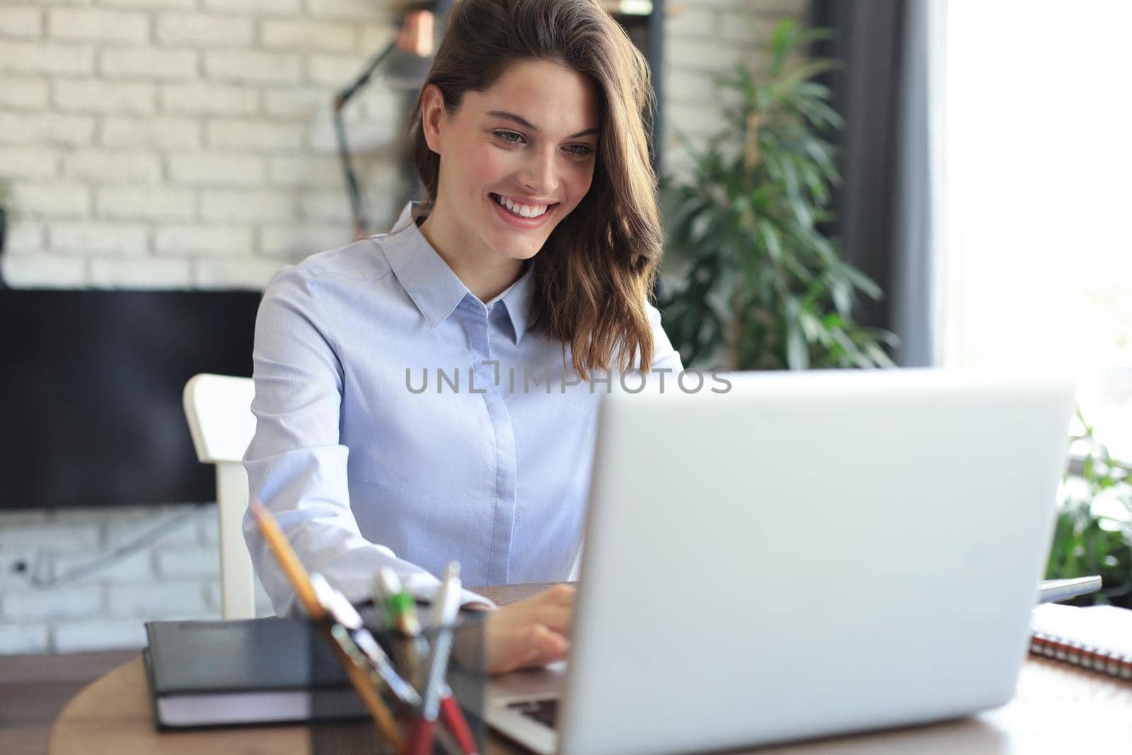 Smiling pretty woman sitting at table, looking at laptop screen. Happy entrepreneur reading message email with good news, chatting with clients online