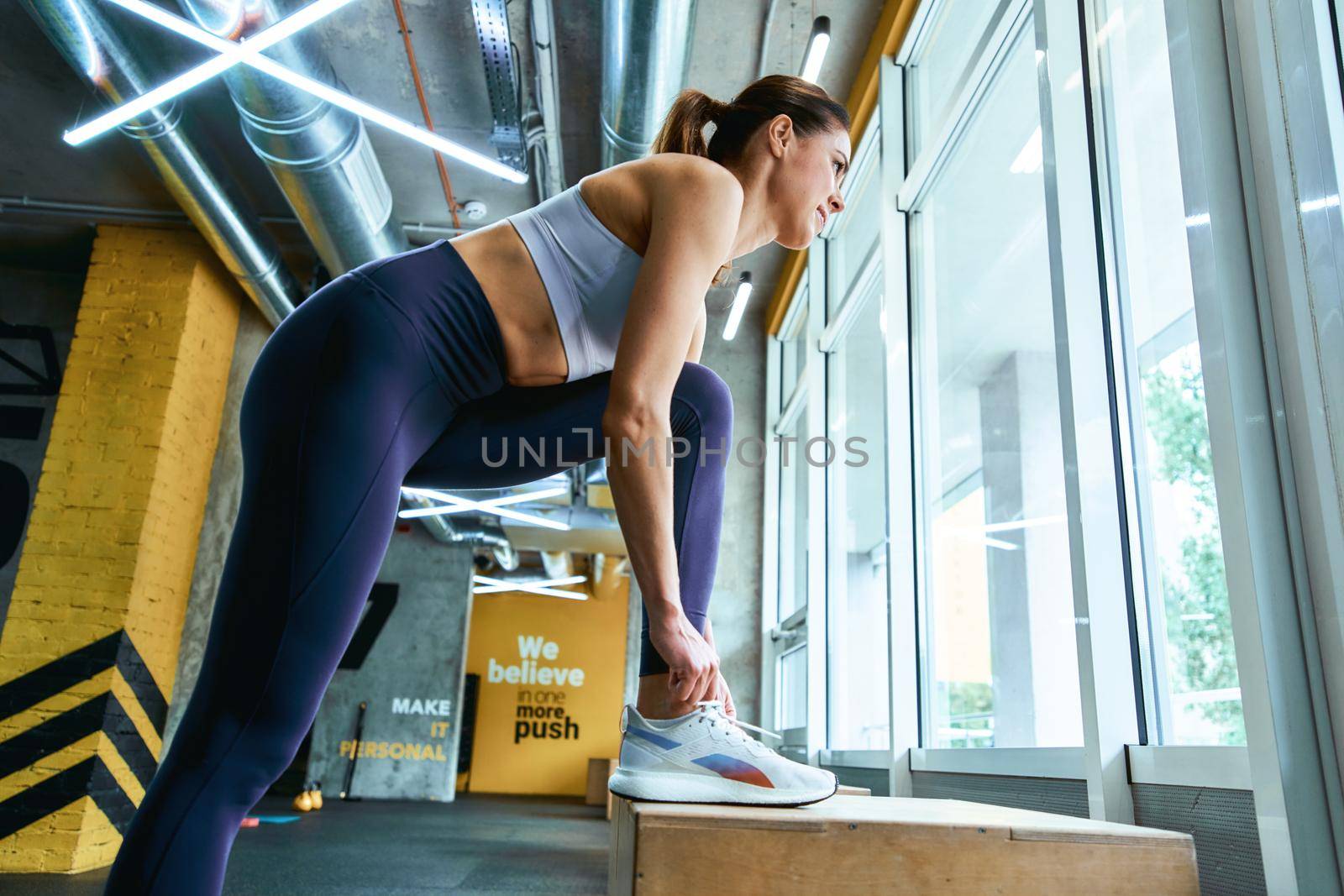 Young beautiful fitness woman in sportswear tying shoelaces and looking at window while exercising at gym. Sport, wellness and healthy lifestyle