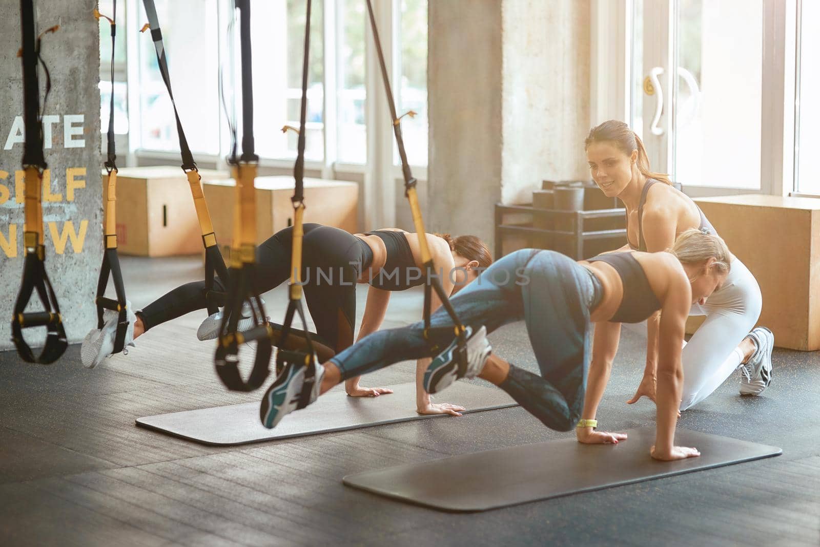 Two young athletic girls exercising with trx fitness straps at industrial gym with assistance of personal trainer by friendsstock