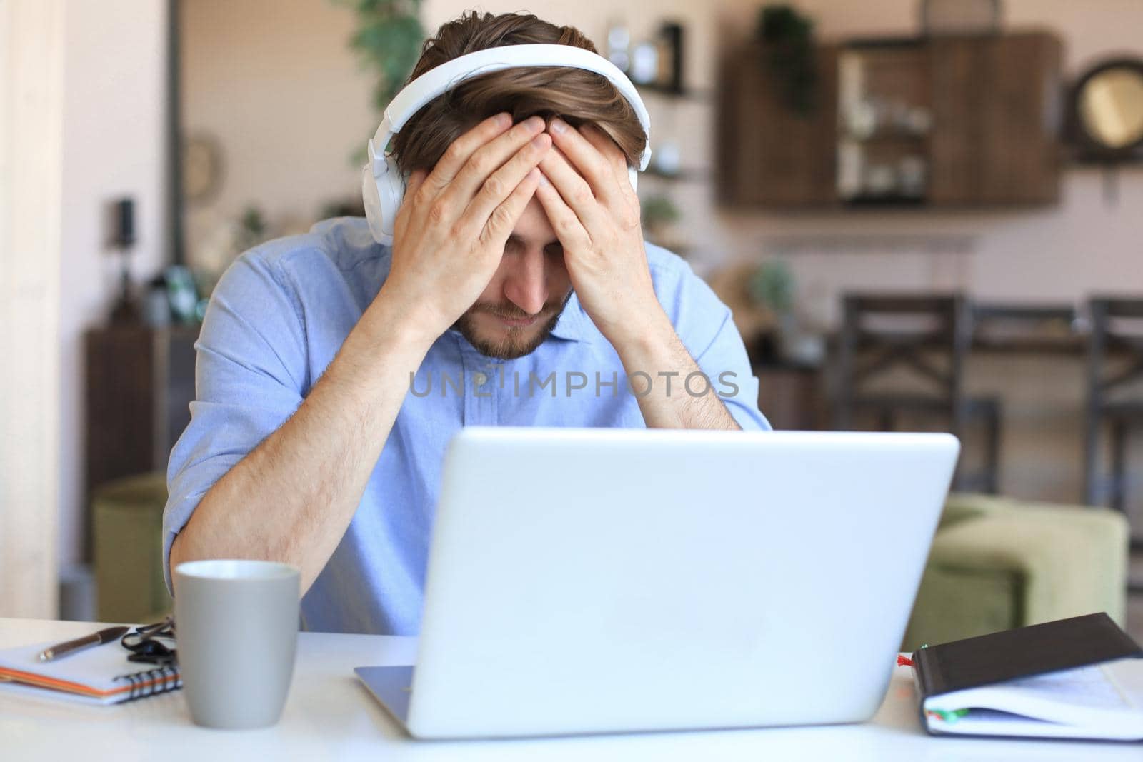 Unhappy frustrated young male holding head by hands sitting with laptop behind desk at home