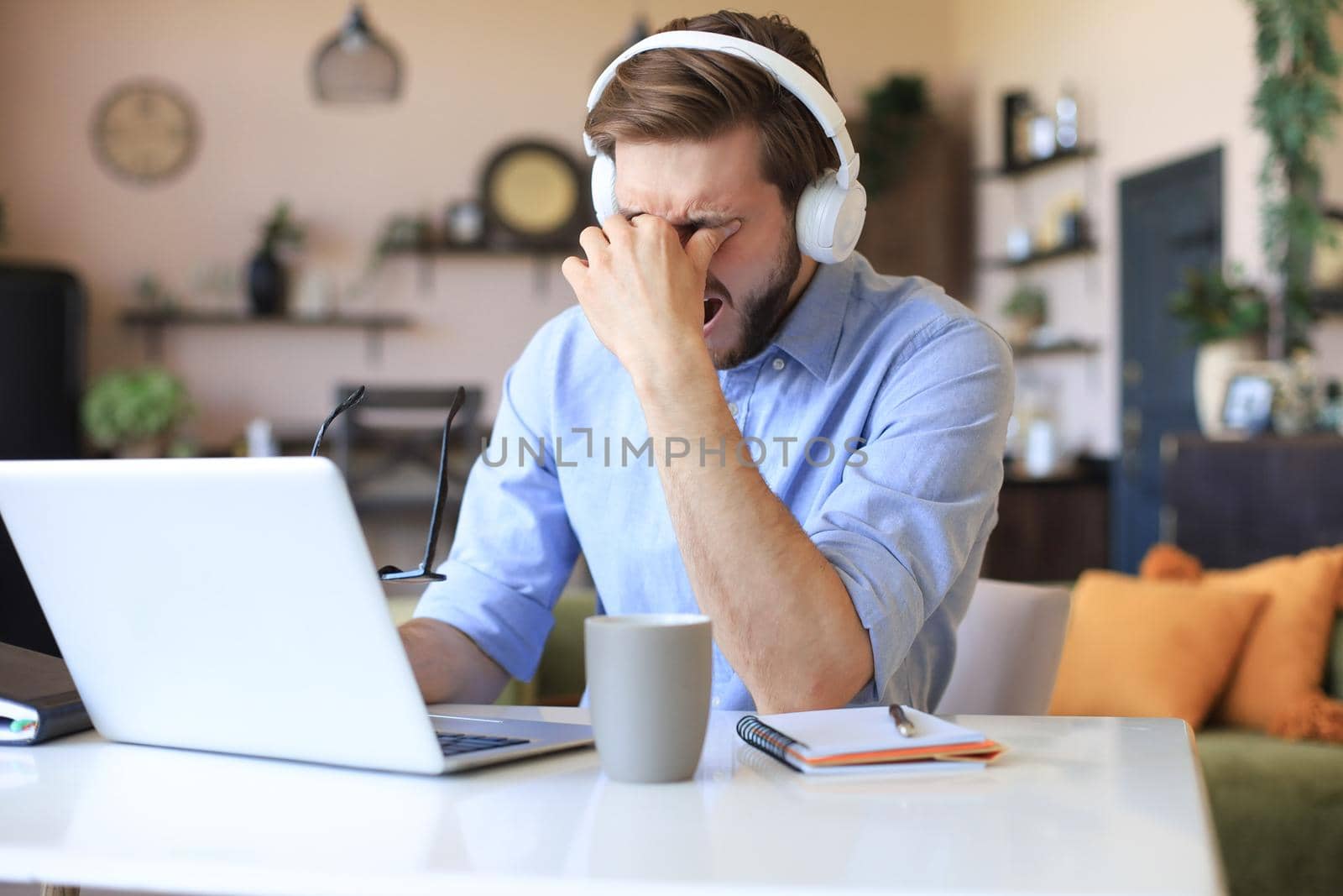 Unhappy frustrated young male holding head by hands sitting with laptop behind desk at home