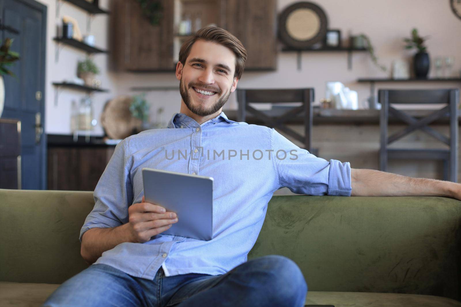 Smiling handsome man holding and using tablet with social networks at home, happy freelancer making online shopping, sitting on cozy sofa at living room