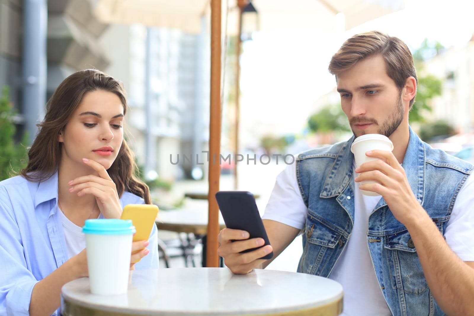 Attractive young couple in love drinking coffee while sitting at the cafe table outdoors, using mobile phone