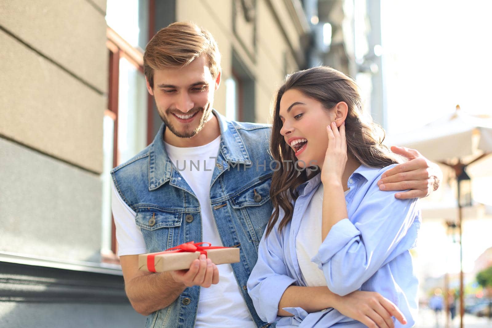 Astonished excited couple in summer clothes smiling and holding present box together while standing on city street