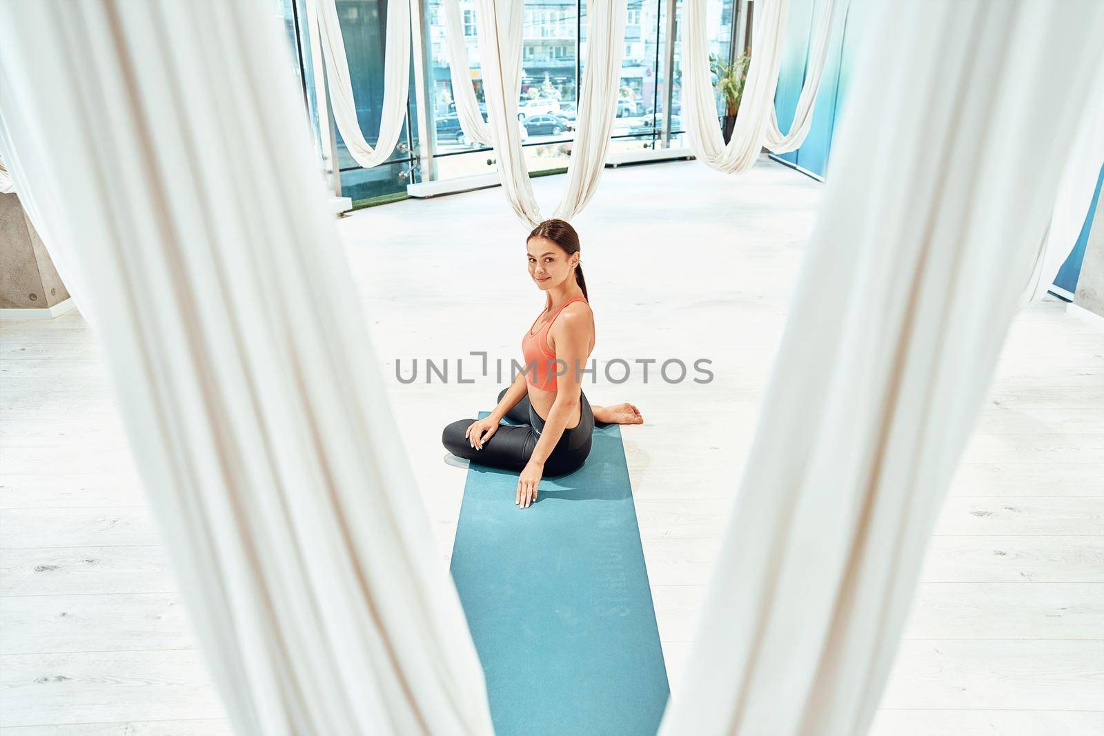 Young beautiful caucasian fitness woman in sportswear sitting on the floor in studio, looking at camera and smiling while practicing fly or aerial yoga. Sport, wellness and healthy lifestyle