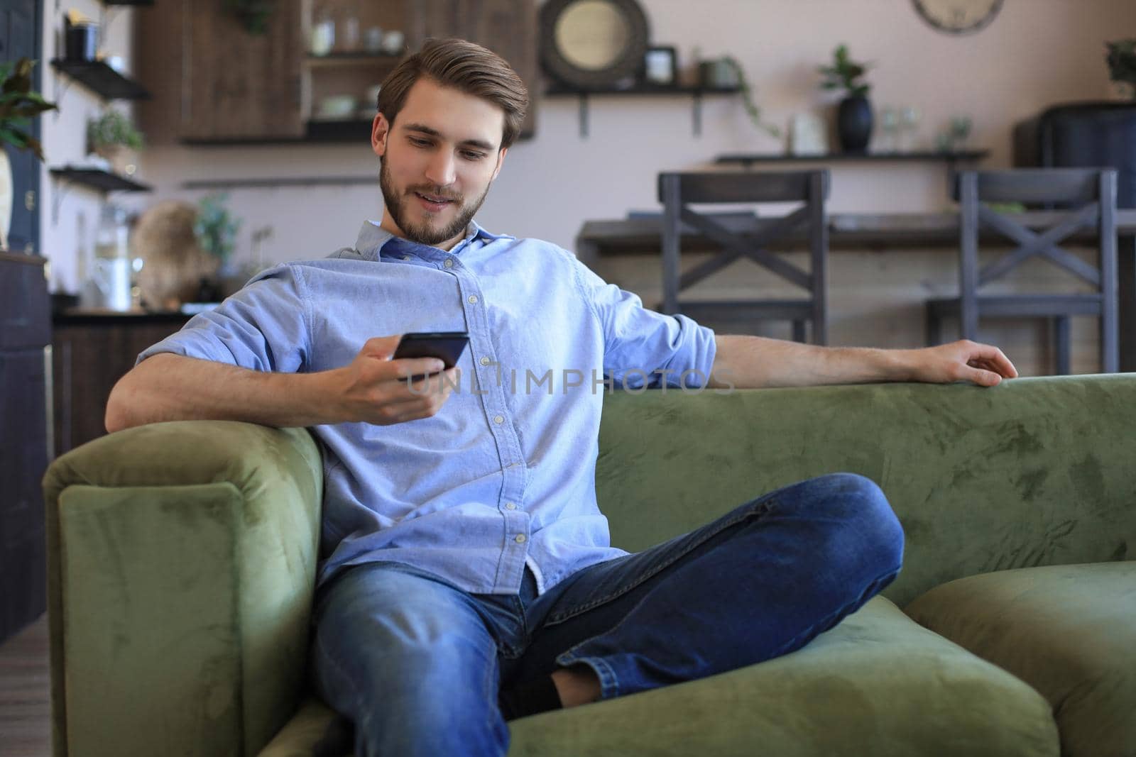 Attractive young man relaxing on a couch at home and using mobile phone for cheking social nets