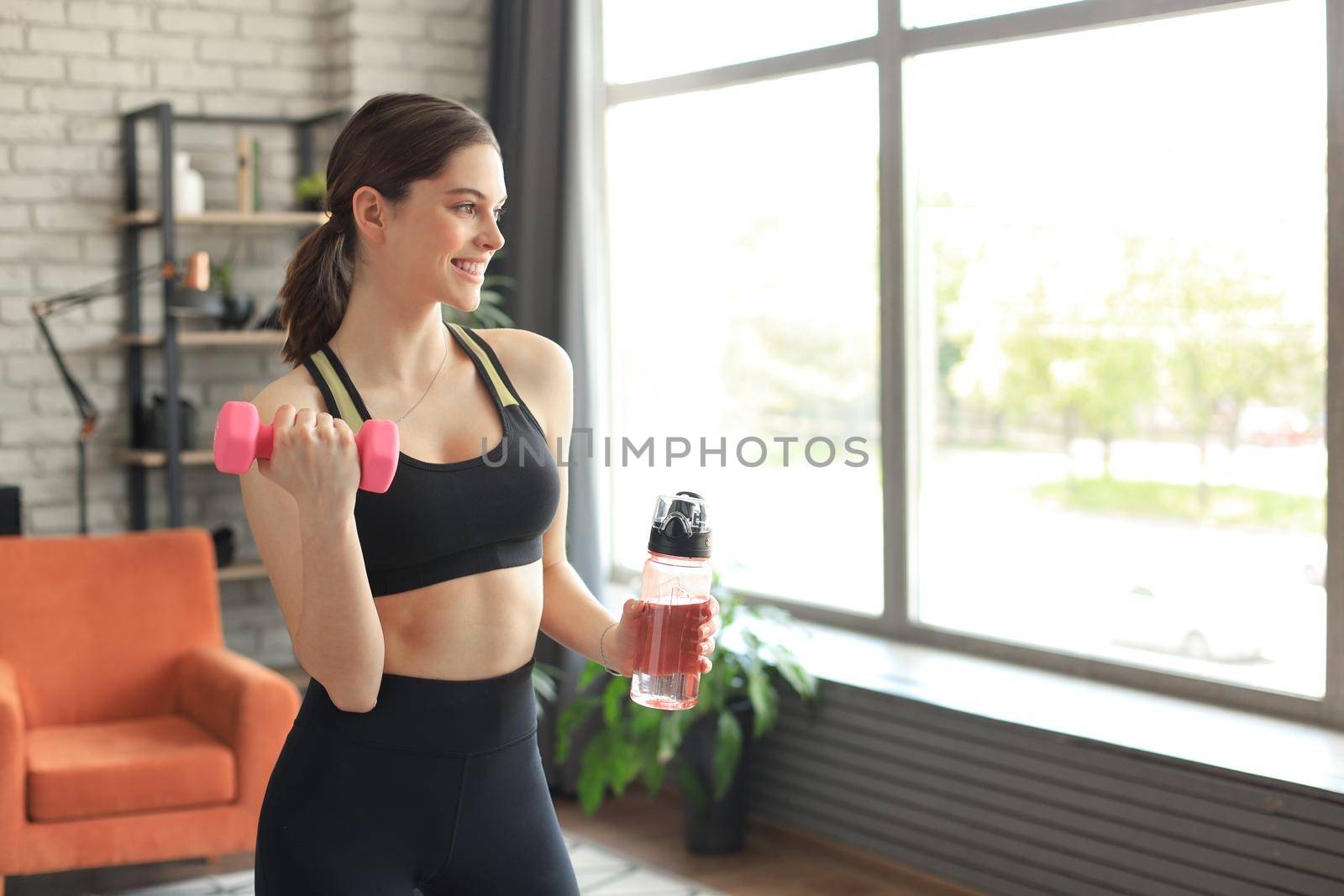 Young beautiful woman standing with dumbbell and drink water after exercising. Attractive female bodybuilder working out. Fitness and healthy lifestyle concept.