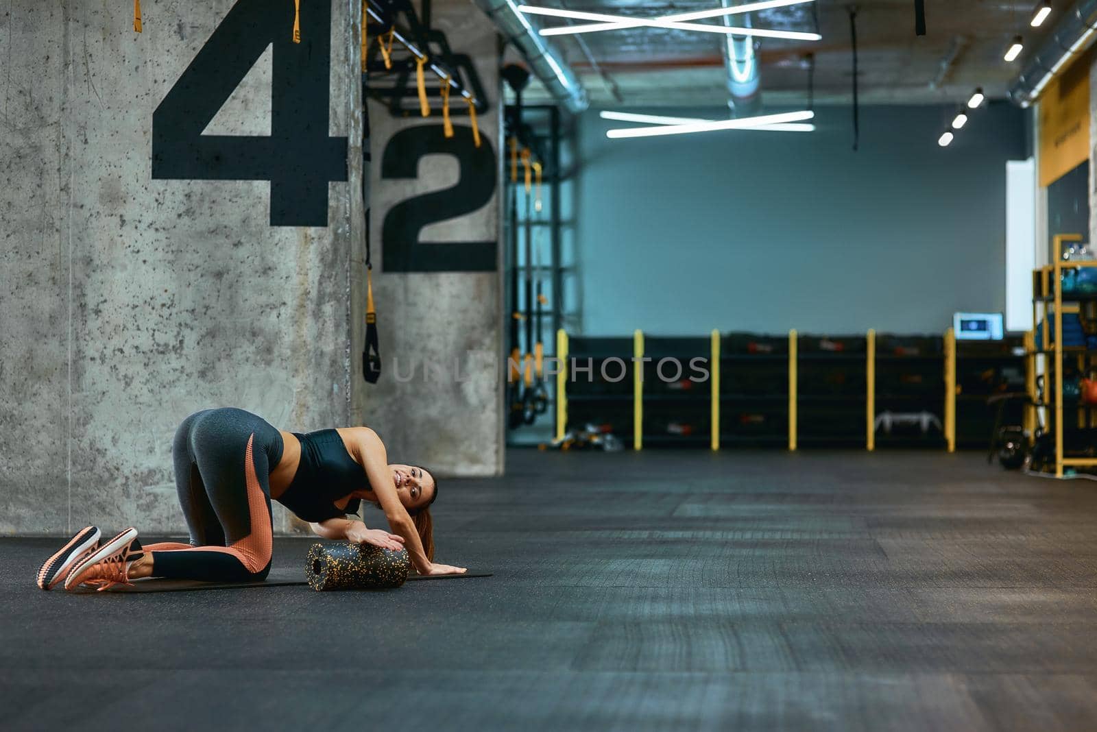 Full length of a young sportive woman in sportswear exercising with foam roller, doing stretching exercises on yoga mat at industrial gym. Sport, training, workout and healthy lifestyle
