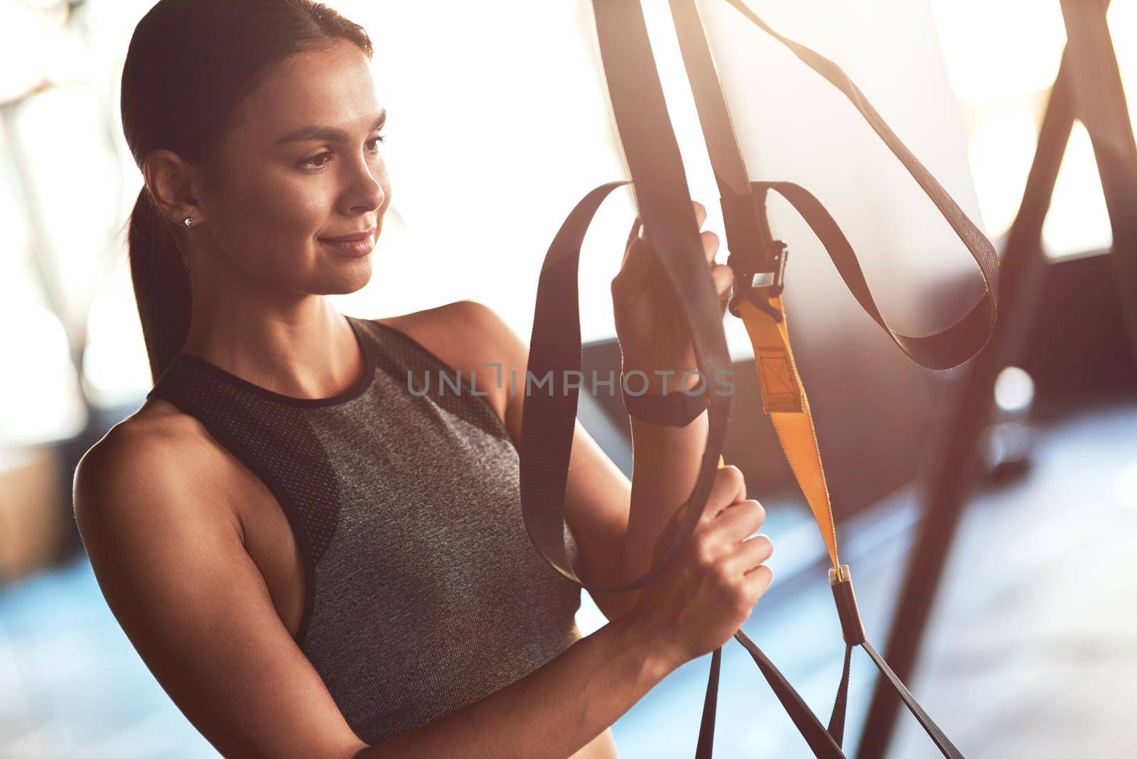 Young beautiful sportive woman adjusting trx fitness straps while exercising at gym by friendsstock