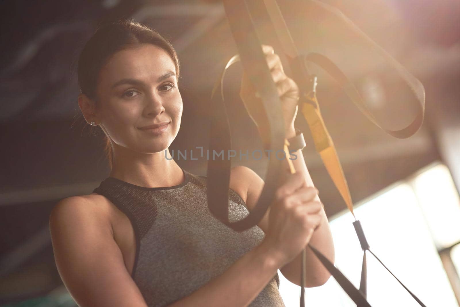 Portrait of a young attractive woman adjusting trx fitness straps, looking at camera and smiling while exercising at gym. Sport, workout, wellness and healthy lifestyle