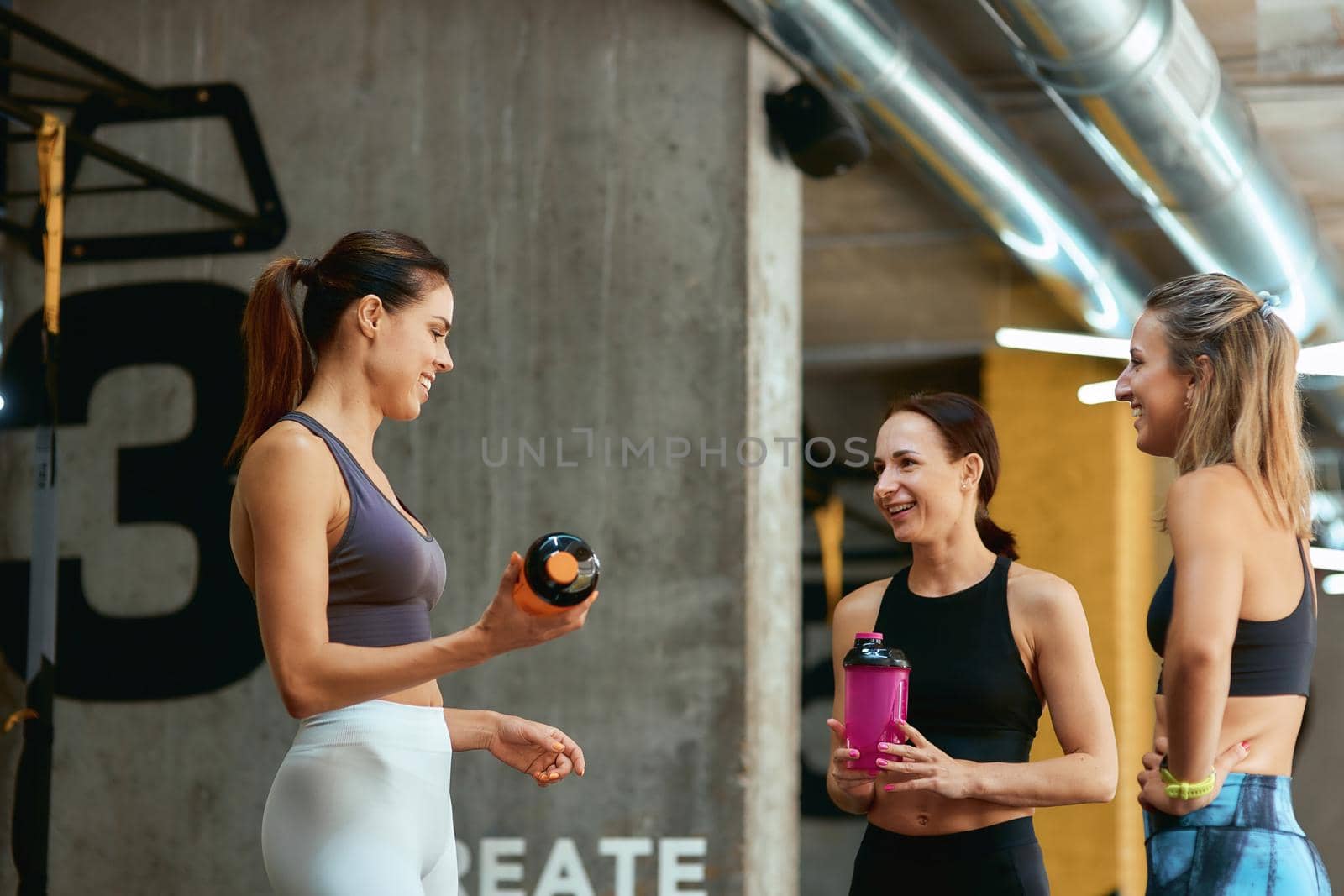 Resting after workout. Group of three young beautiful fitness women in sportswear talking and smiling while taking a break at gym. Sport, wellness and healthy lifestyle