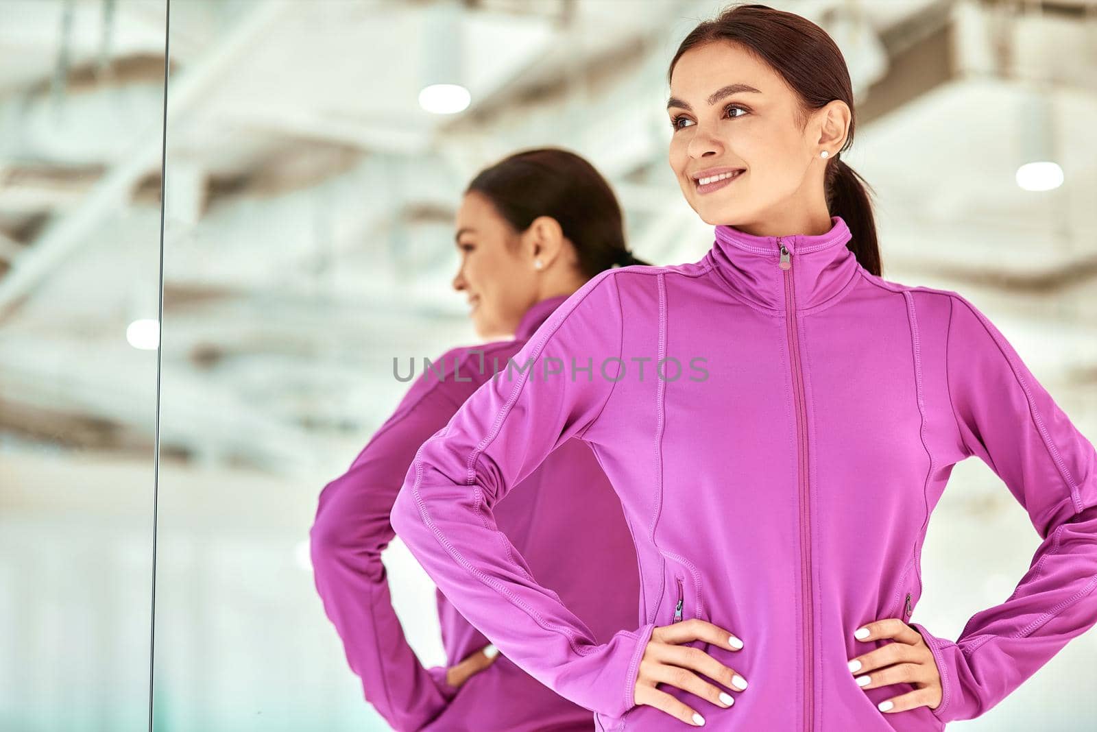 Young beautiful woman, female fitness instructor in sportswear keeping arms on hips, looking away and smiling while standing in studio or gym. Sport, workout, wellness and healthy lifestyle