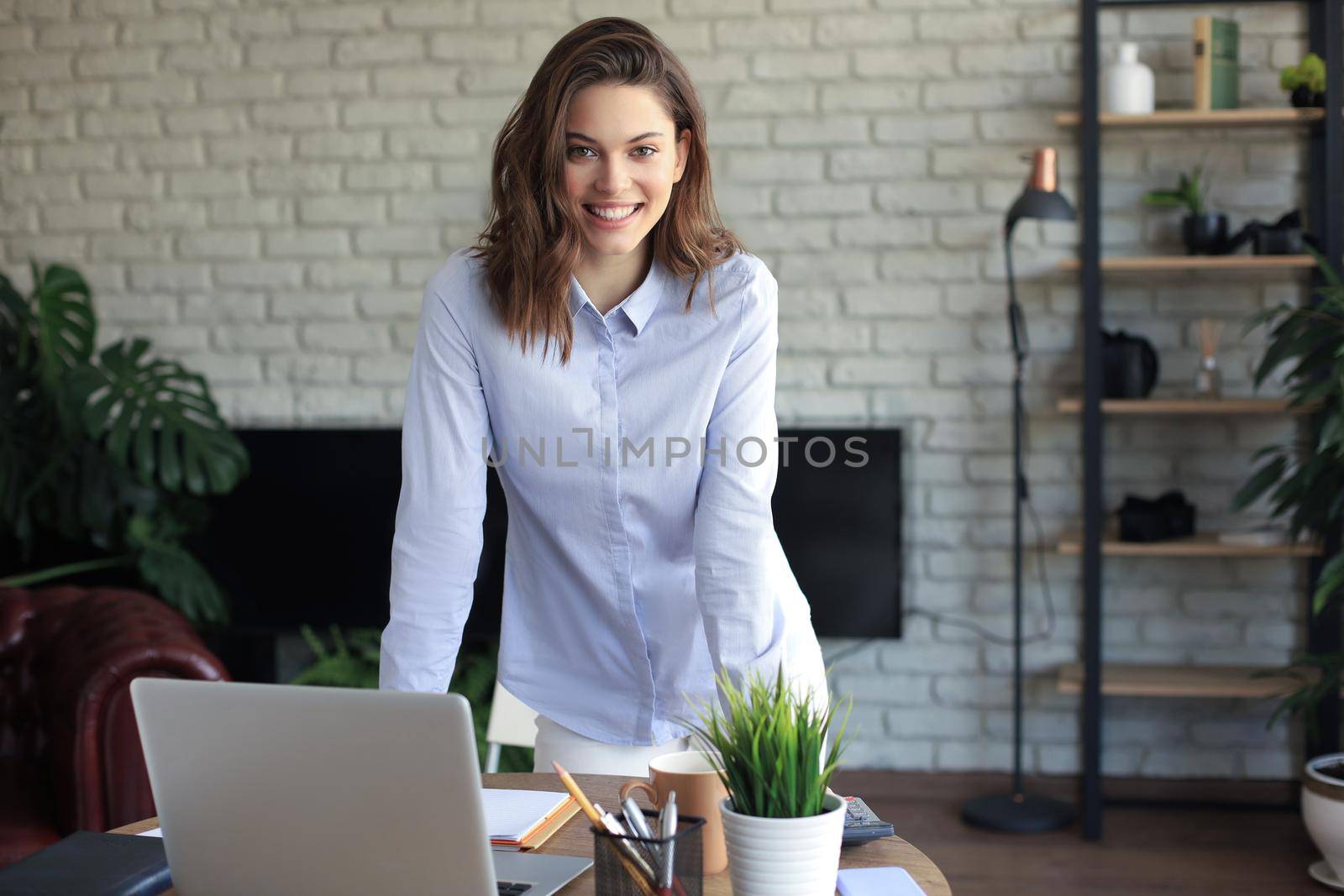 Confident young businesswoman with a friendly smile standing behind her desk in a home office looking at the camera