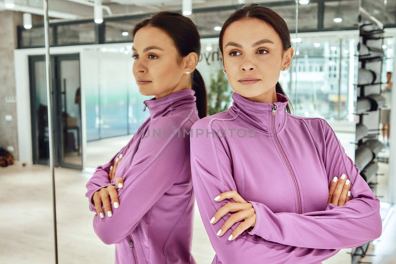 Fitness instructor. Portrait of a young beautiful woman in sportswear standing in studio or gym against mirror wall and looking away. Sport, wellness and healthy lifestyle