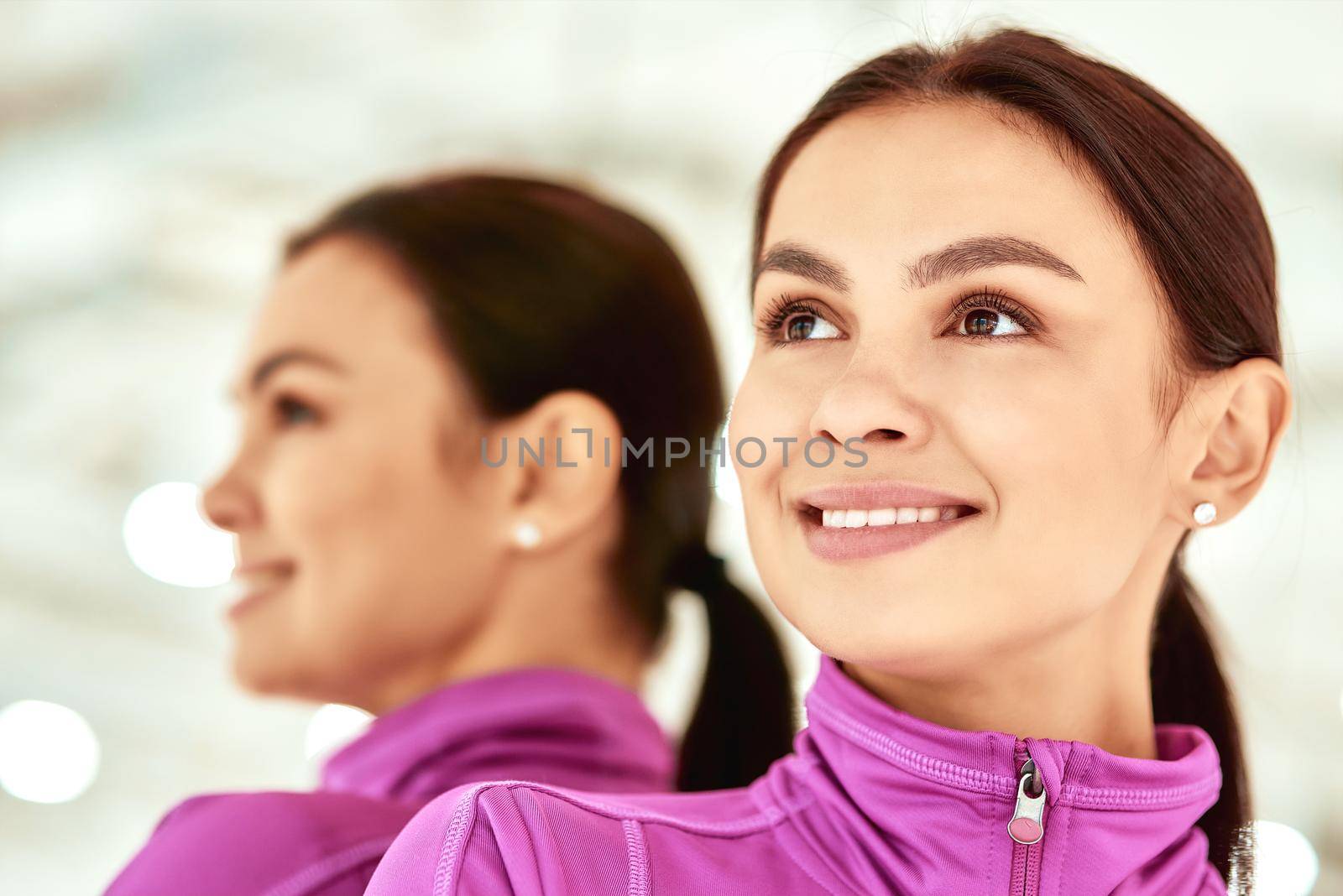 Close up portrait of a young beautiful caucasian woman, female fitness instructor in sportswear looking away and smiling standing in studio or gym against mirror by friendsstock