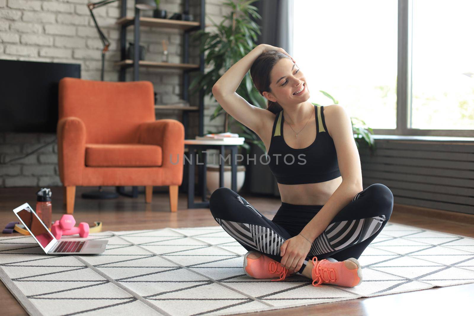Fitness beautiful slim woman is sitting on the floor with dumbbells and bottle of water using laptop at home in the living room. Stay at home activities. by tsyhun