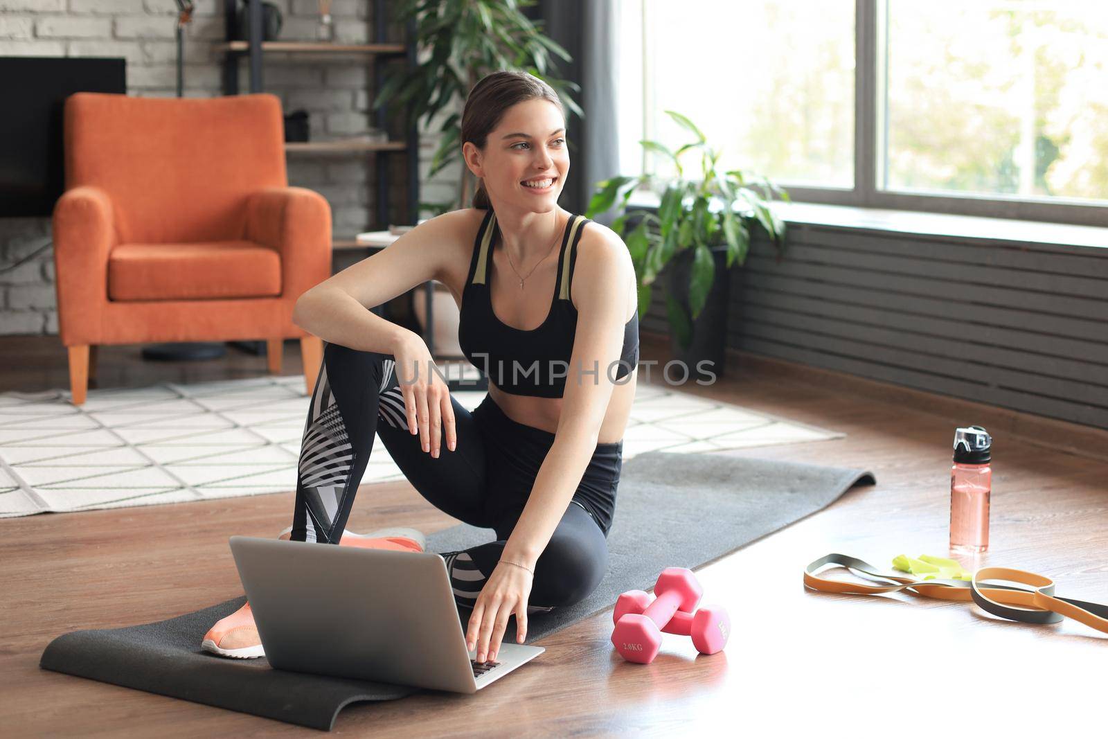 Beautiful slim sporty woman in sportswear is sitting on the floor with dumbbells and bottle of water and is using a laptop at home in the living room. Healthy lifestyle. Stay at home activities. by tsyhun