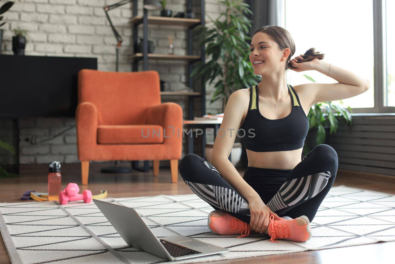 Fitness beautiful slim woman is sitting on the floor with dumbbells and bottle of water using laptop at home in the living room. Stay at home activities