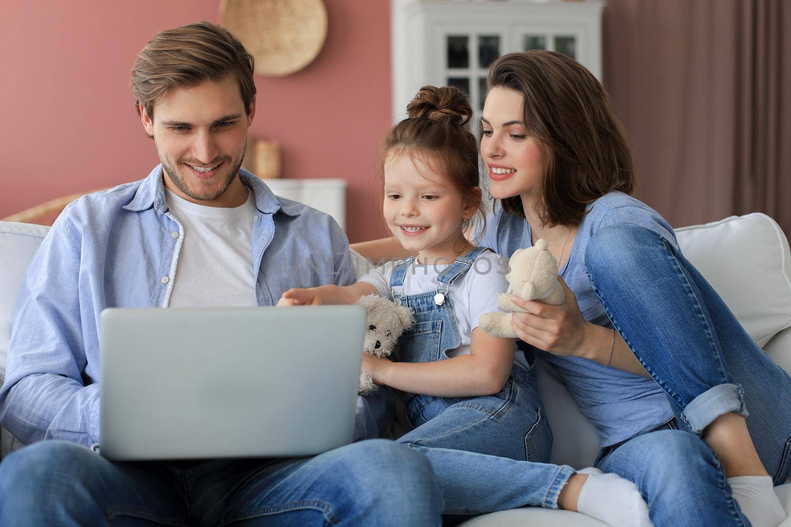 Positive friendly young parents with smiling little daughter sitting on sofa together answering video call on laptop and waving hand in greeting while relaxing at home on weekend