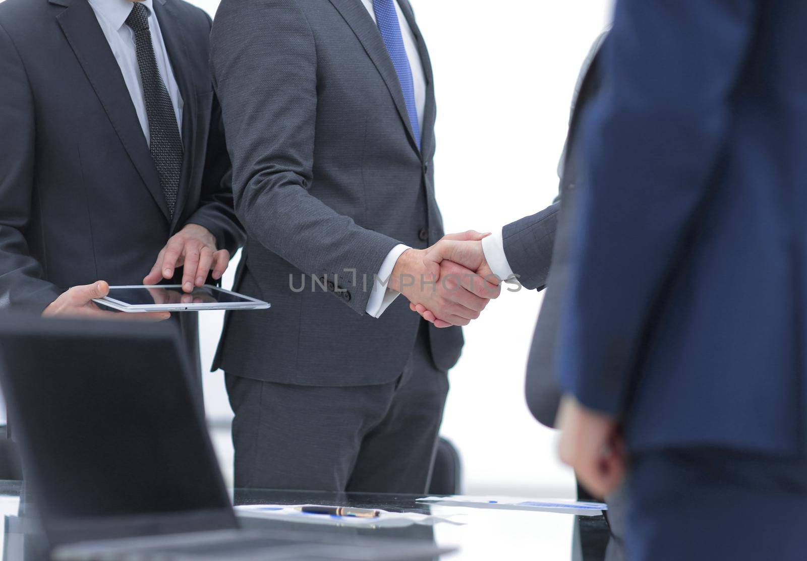 Two cheerful businessmen shaking hands with smile at office with their coworkers.