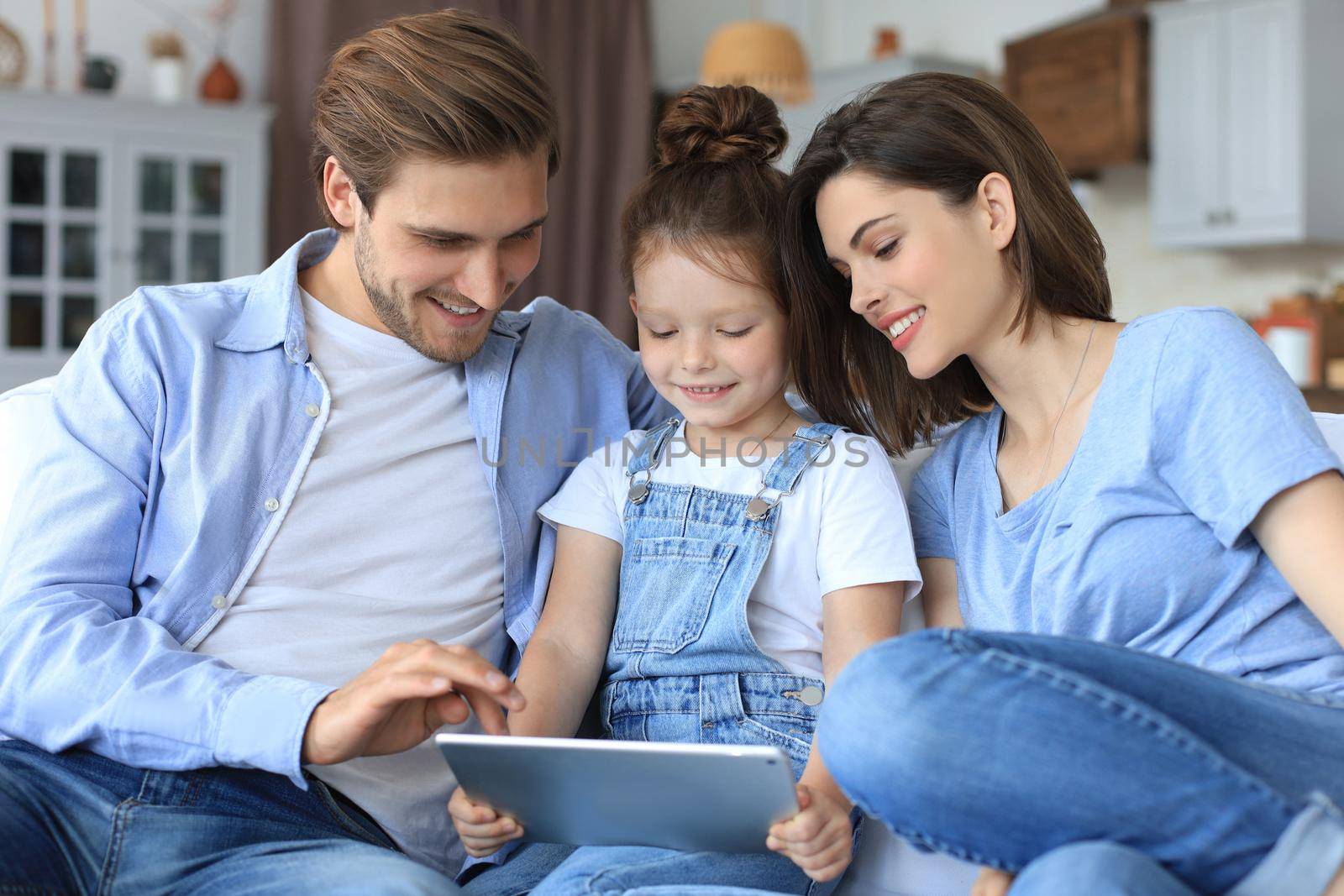 Positive friendly young parents with smiling little daughter sitting on sofa together answering video call on digital tablet while relaxing at home on weekend