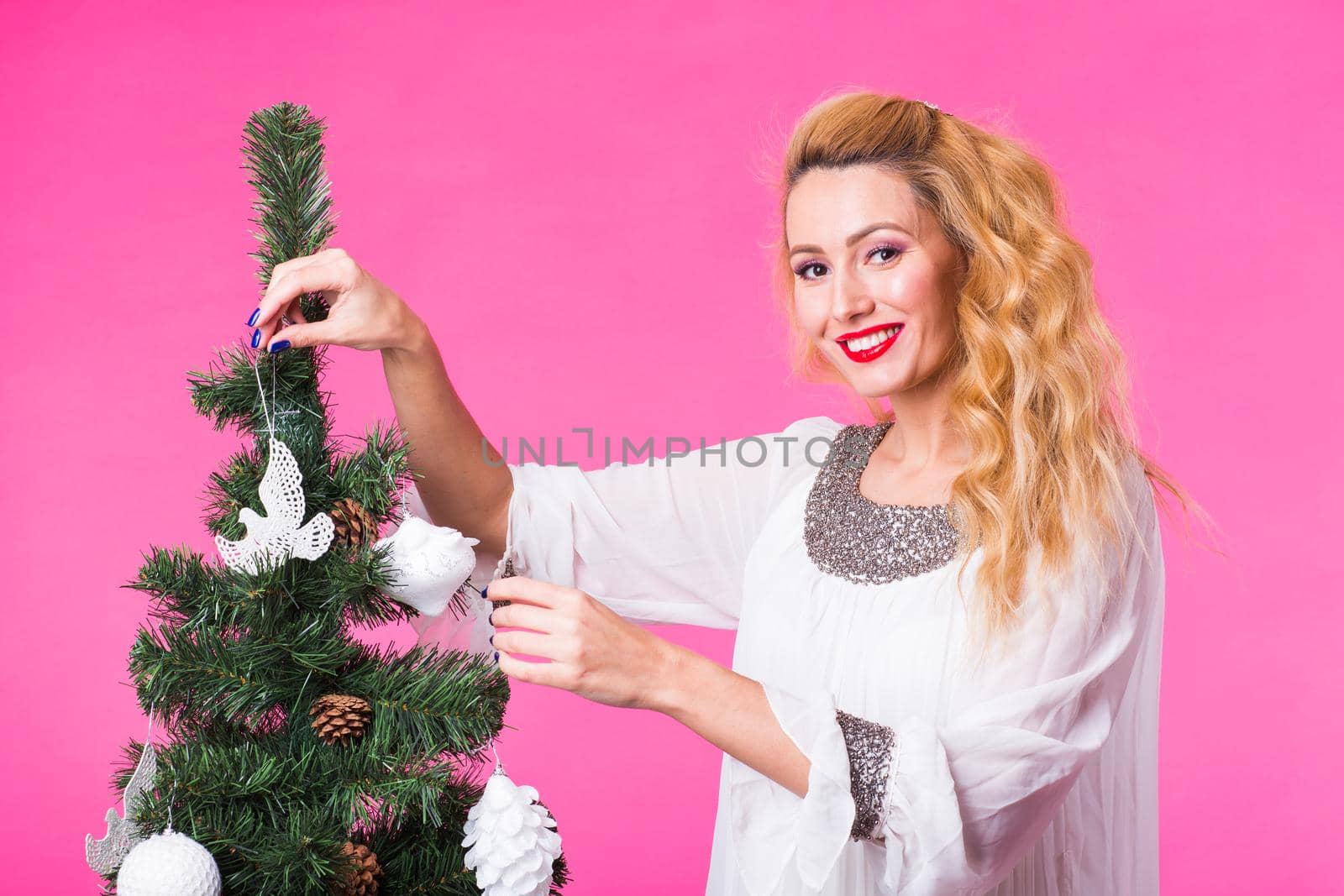 Christmas, holidays and people concept - young happy blonde woman decorating christmas tree on pink background by Satura86