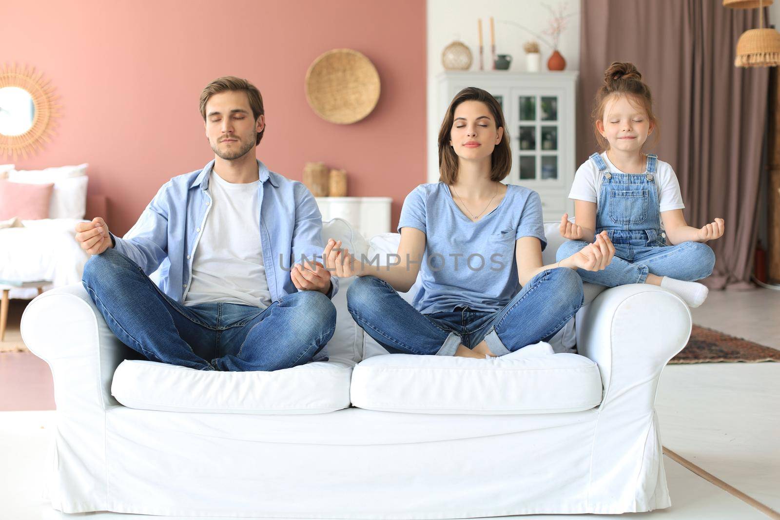 Calm young family with little daughter sit on couch practice yoga together, happy parents with small preschooler girl child rest on sofa meditate relieve negative emotions on weekend at home