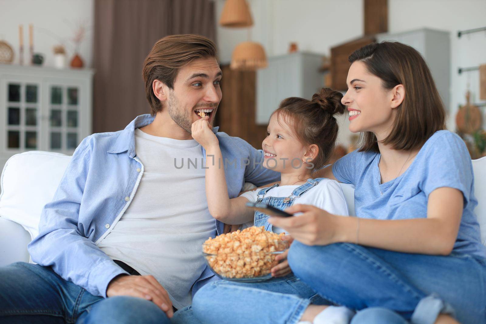 Happy family with child sitting on sofa watching tv and eating popcorn, young parents embracing daughter relaxing on couch together