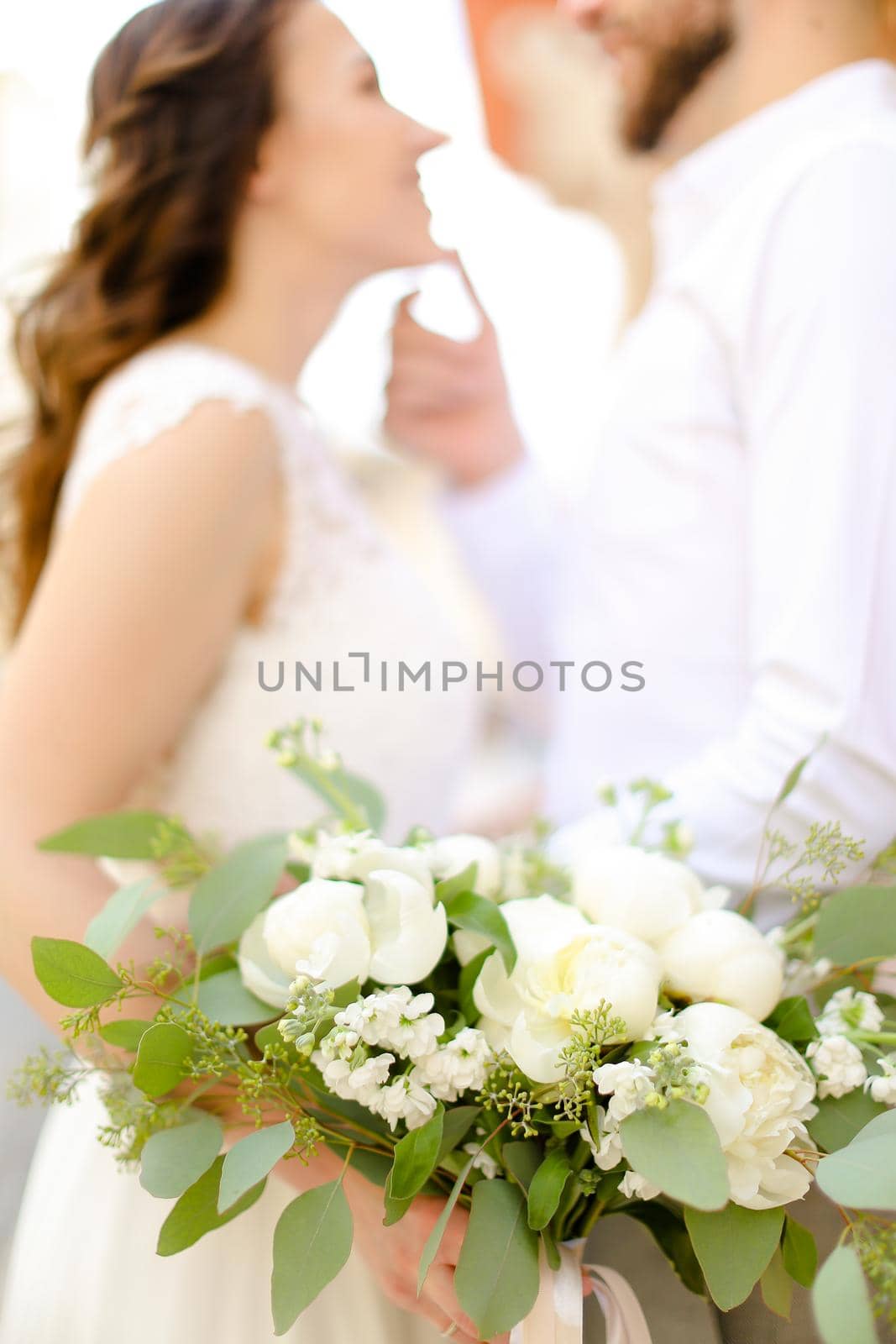 Happy groom hugging beautiful bride keeping flowers and wearing white dress. Concept of married couple and love, wedding photo session.