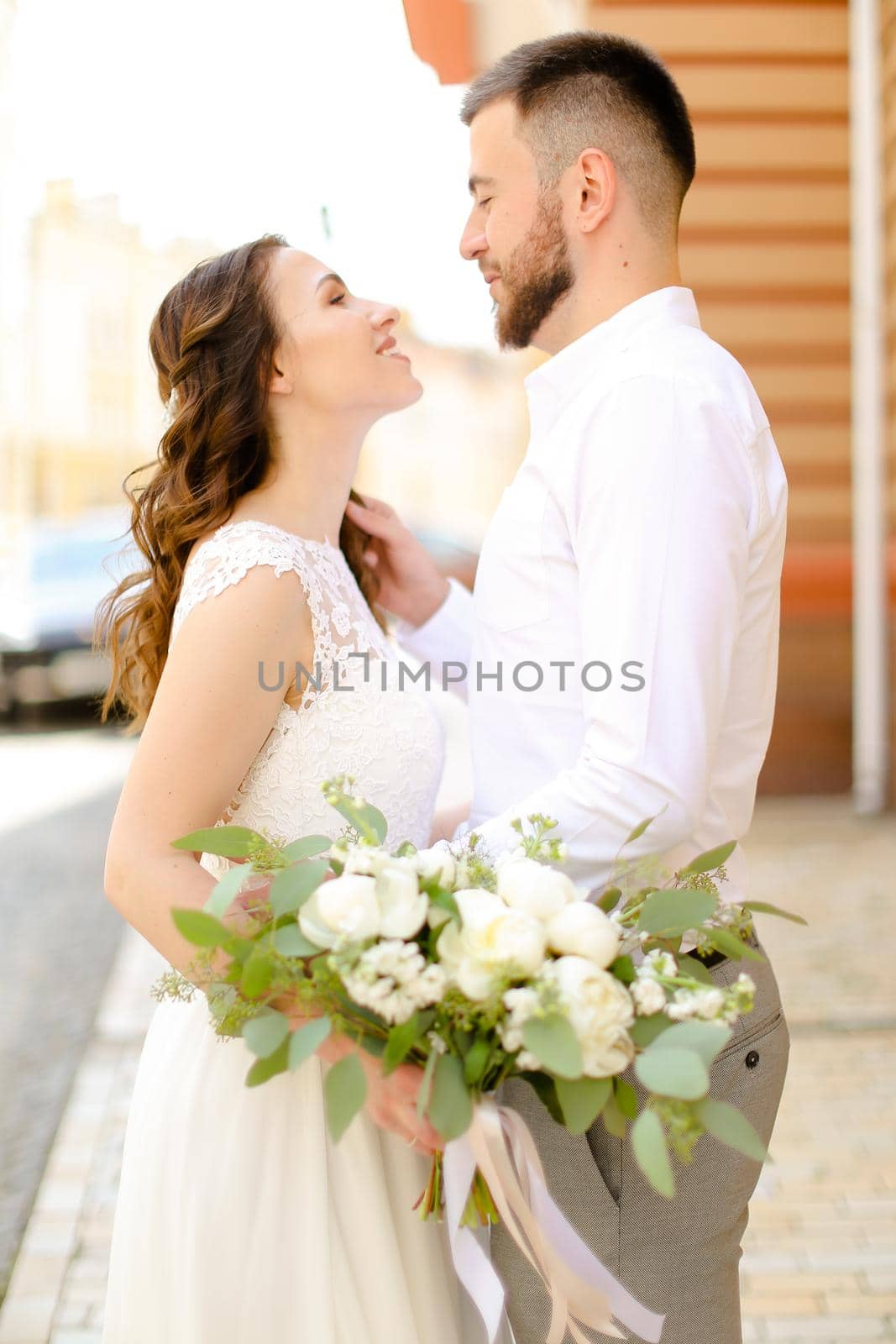 Happy handsome groom hugging bride keeping flowers and wearing white dress. Concept of married couple and love, wedding photo session.