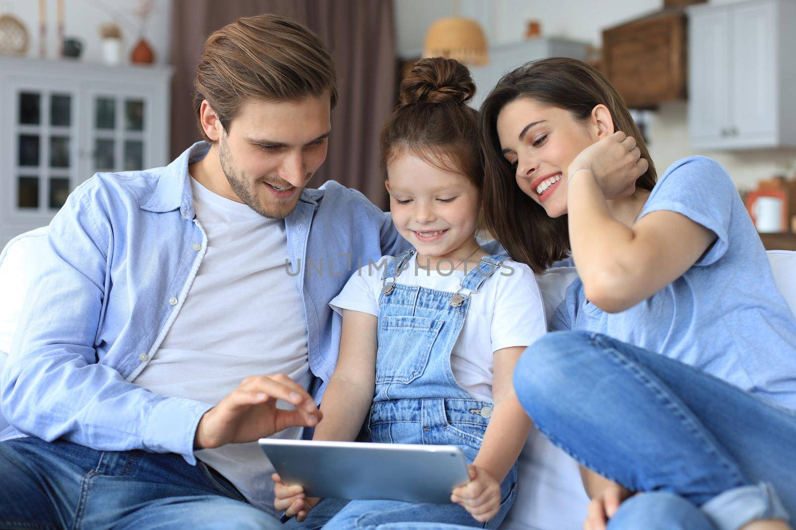 Positive friendly young parents with smiling little daughter sitting on sofa together answering video call on digital tablet while relaxing at home on weekend