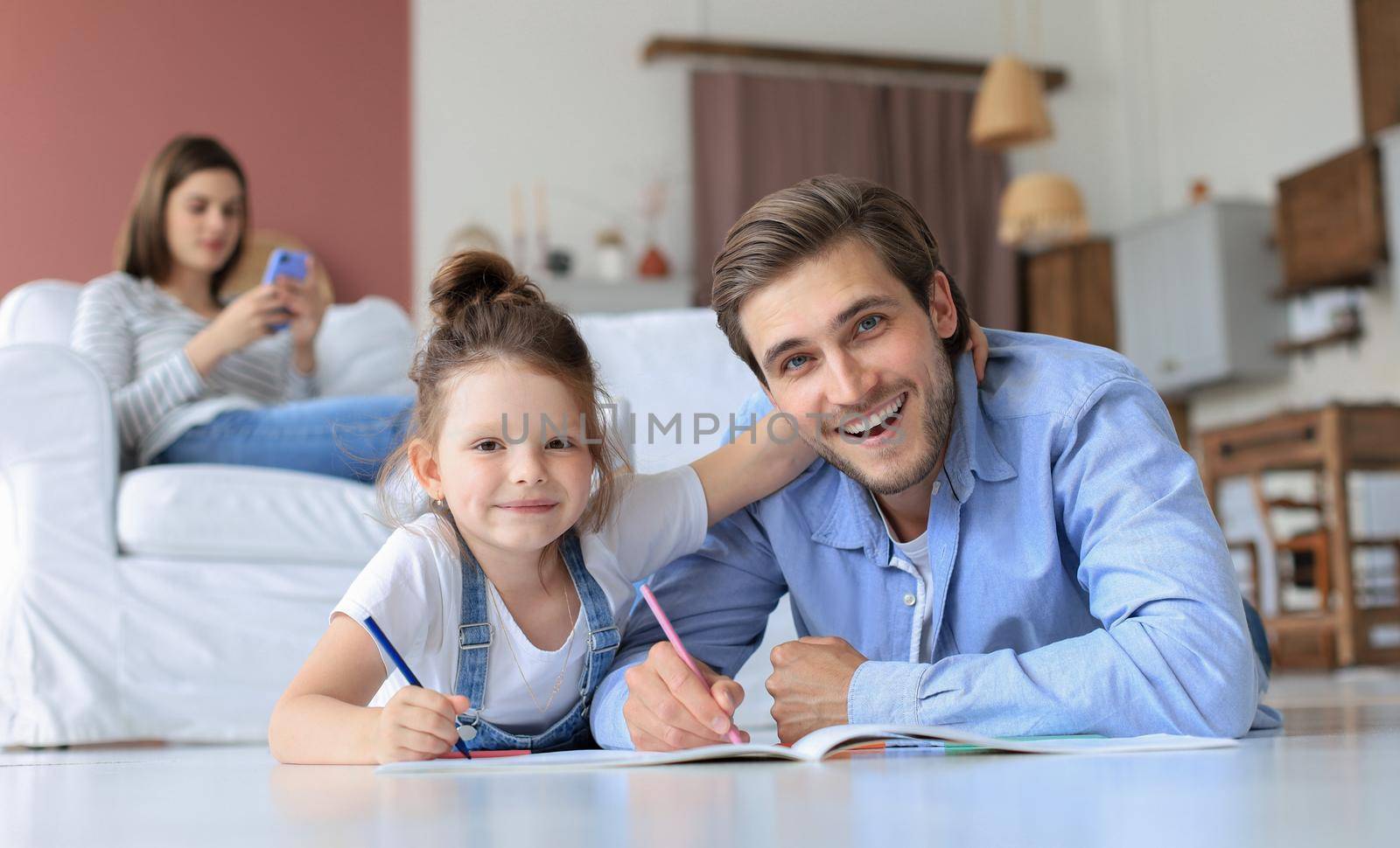 Happy father smilling daughter lying on warm floor enjoying creative activity, drawing pencils coloring pictures in albums, mother resting on couch, family spend free time together