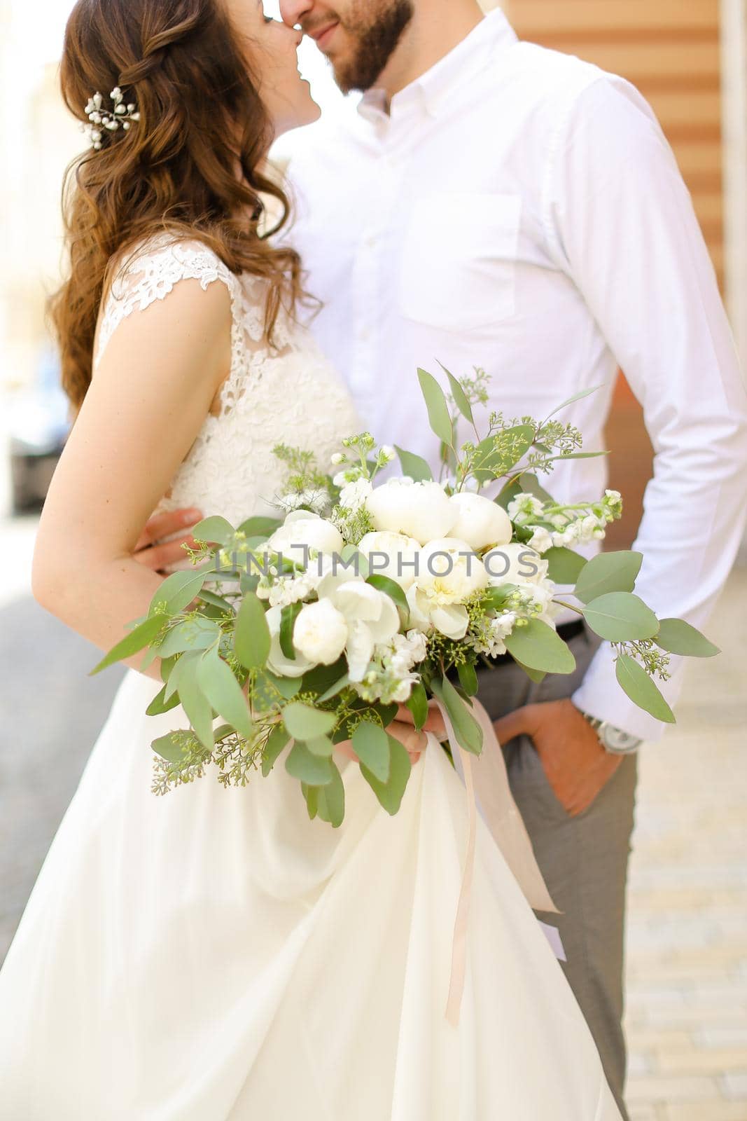 Happy caucasian groom hugging bride keeping flowers and wearing white dress. Concept of married couple and love, wedding photo session.
