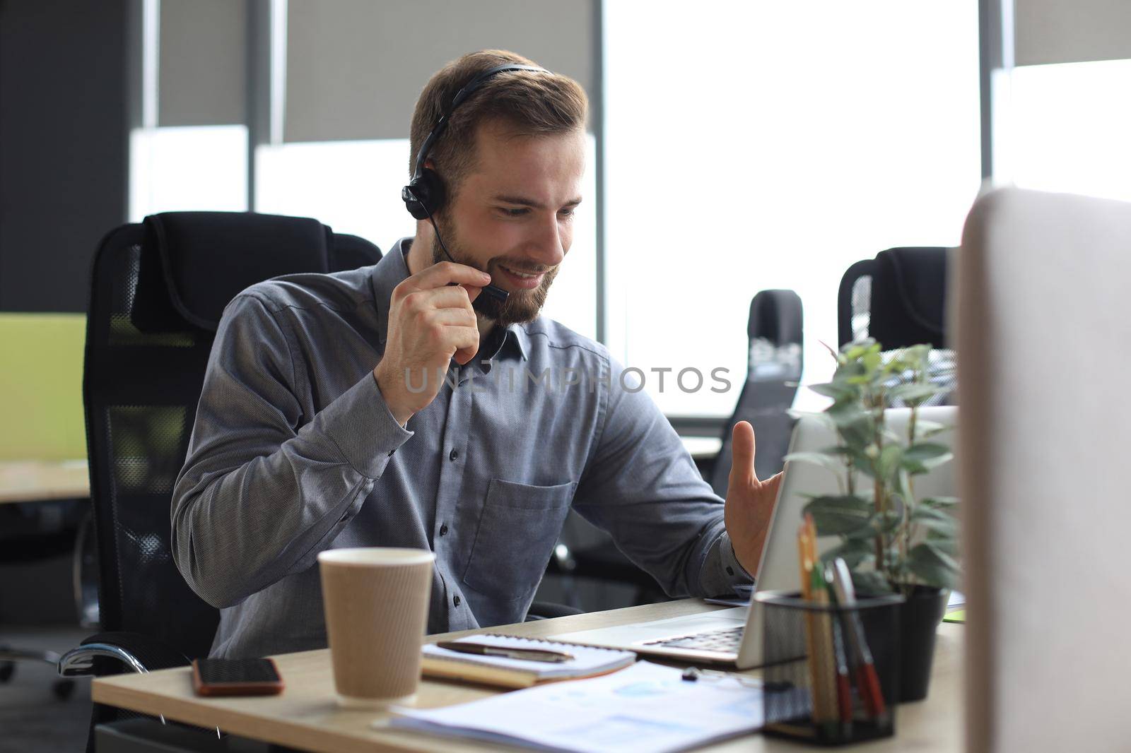 Smiling male business consultant with headphones sitting at modern office, video call looking at laptop screen. Man customer service support agent helpline talking online chat